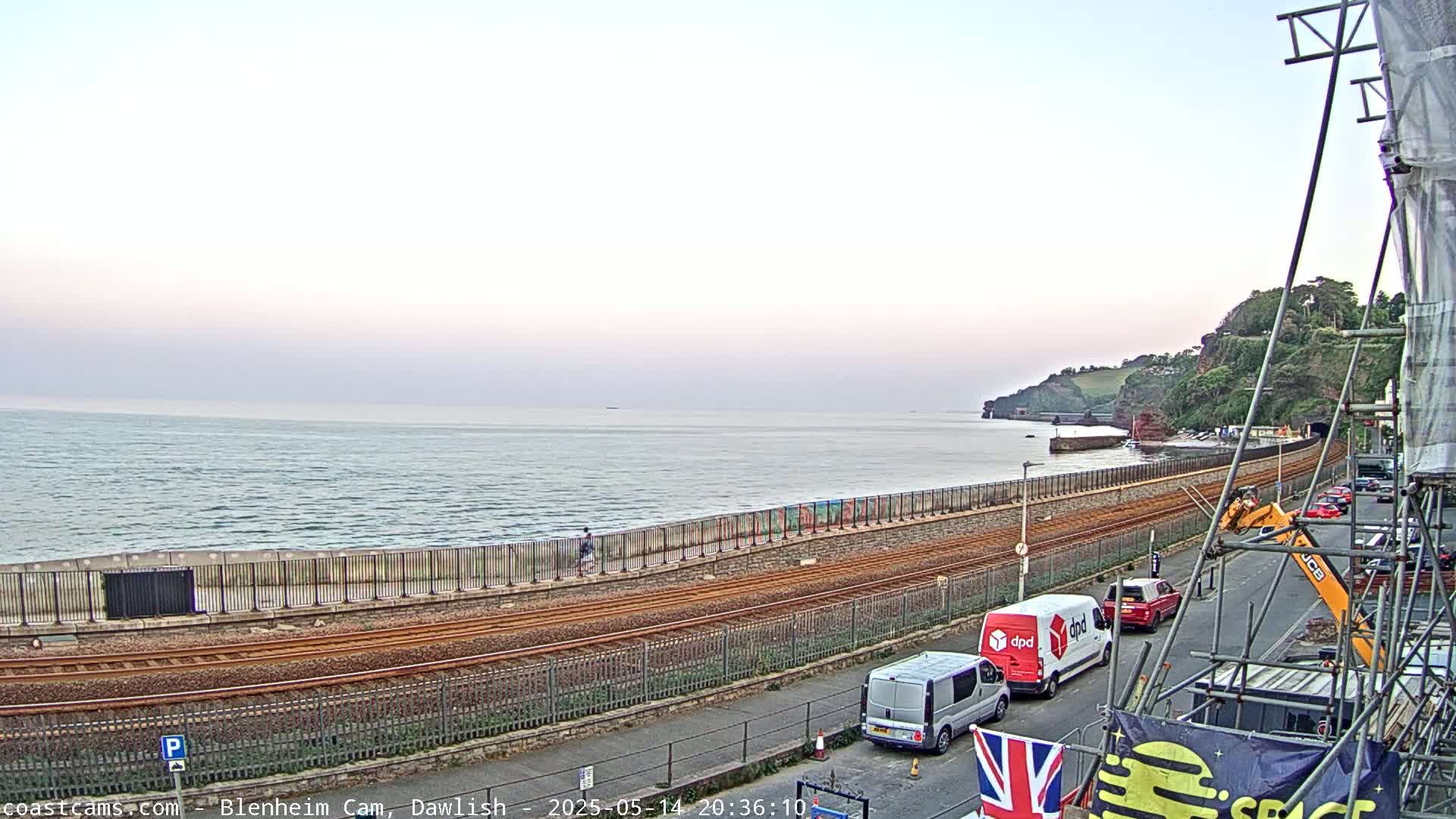 A calm ocean stretches to a coastline featuring train tracks, a seawall, and a road with several vehicles parked, all under a pale sky.