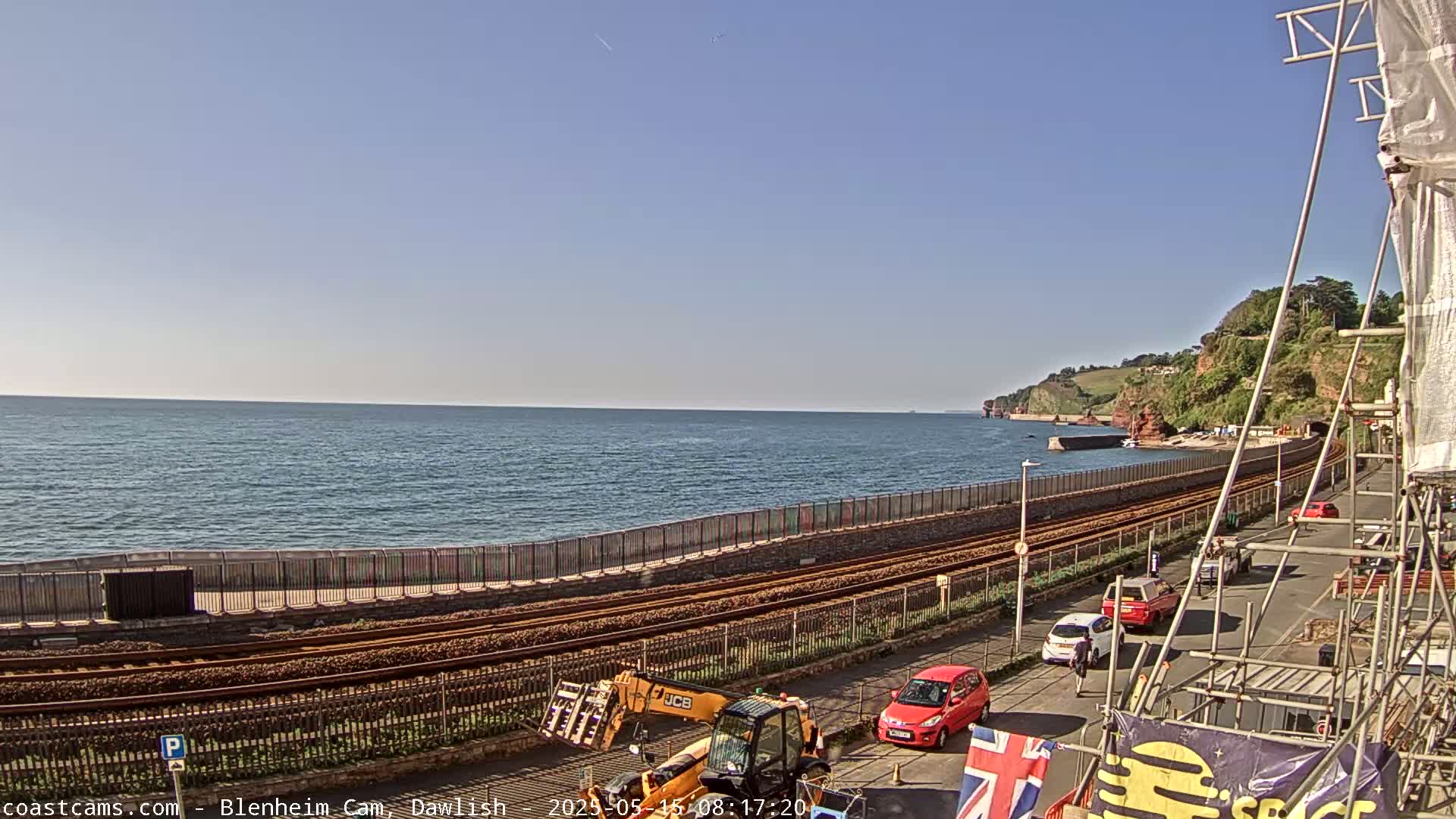 A clear, sunny day reveals a coastal scene with railway tracks running alongside the ocean, construction equipment, and several parked cars near a seaside wall.