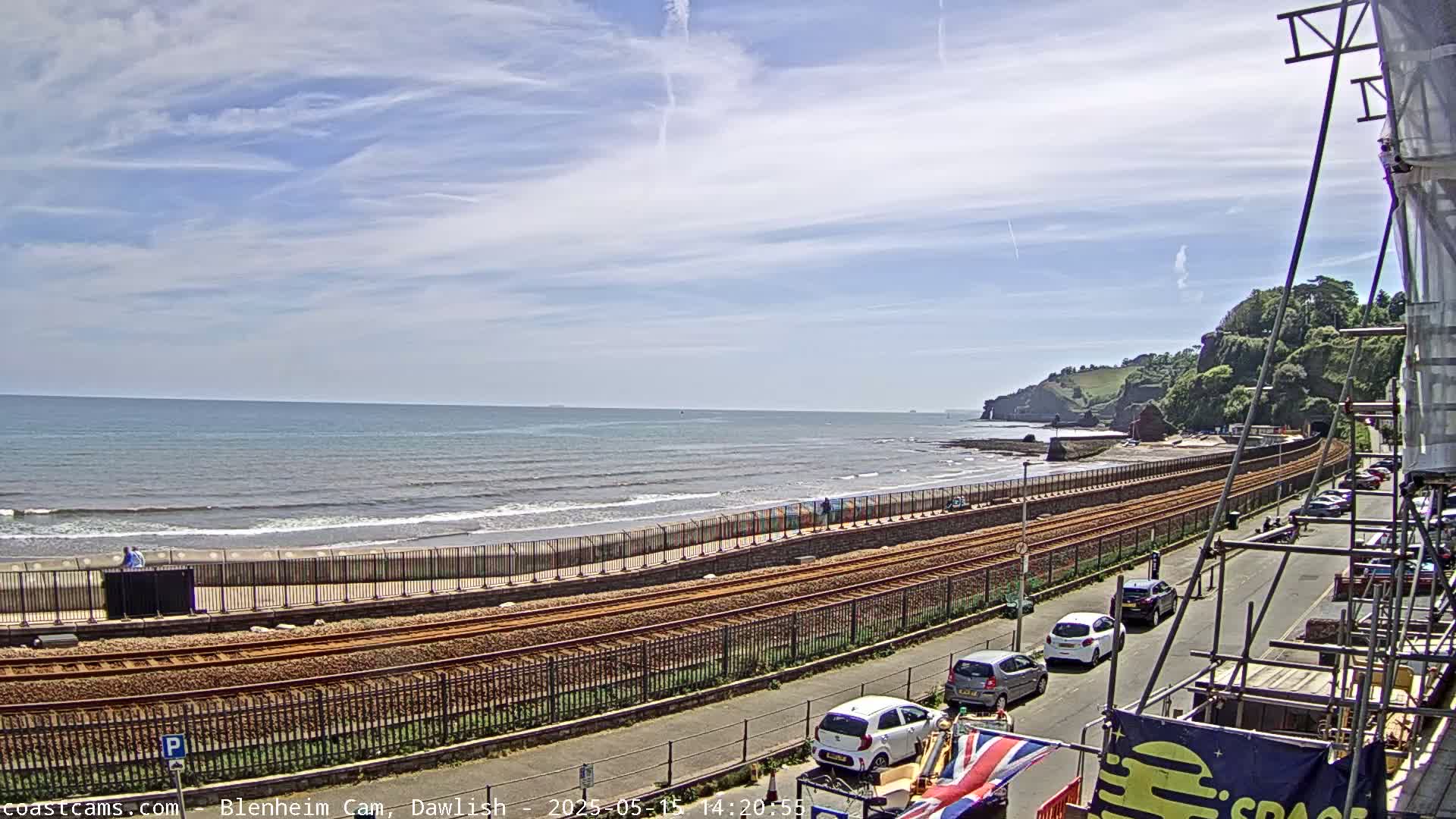 A partly sunny day shows a beach with gentle waves, a railway line running alongside it, a road with parked cars, and green cliffs in the background.