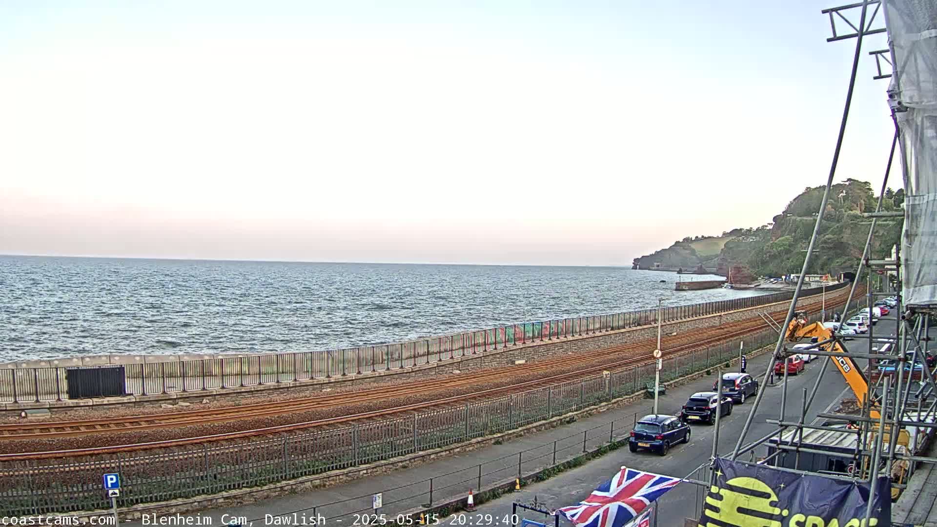 A seaside view shows a calm sea, train tracks running along a seawall, a road with parked cars, and construction scaffolding under a pale sky.