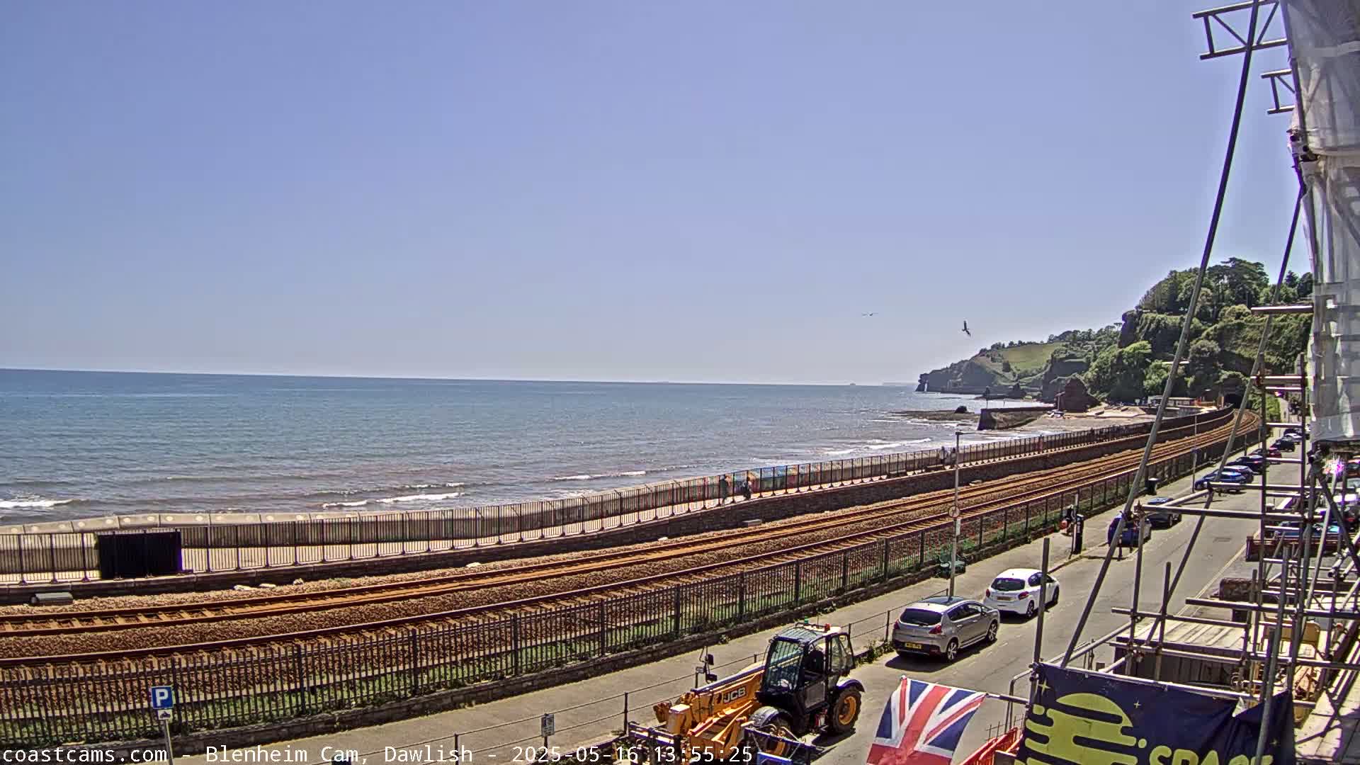 A sunny day shows a beach with a calm ocean, train tracks running alongside a paved road with parked cars, and construction scaffolding visible in the foreground.