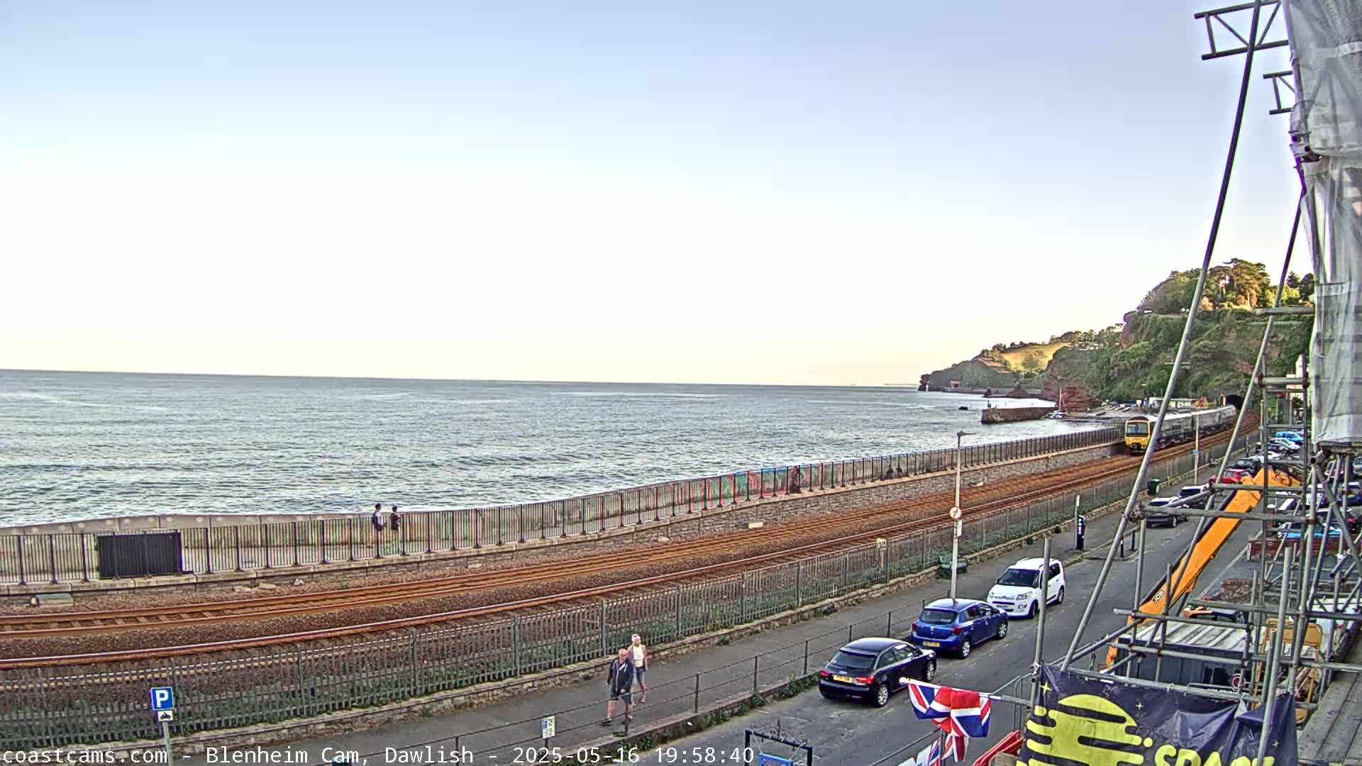 A train passes along a coastal railway line next to a calm sea under a clear sky, with cars parked along a road nearby and several people walking along a seawall.