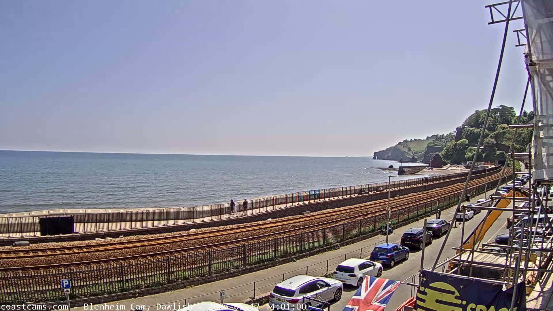 A seaside view on a sunny day shows a calm ocean, a railway track running along a sea wall, a road with parked cars, and some greenery on a cliff in the background.