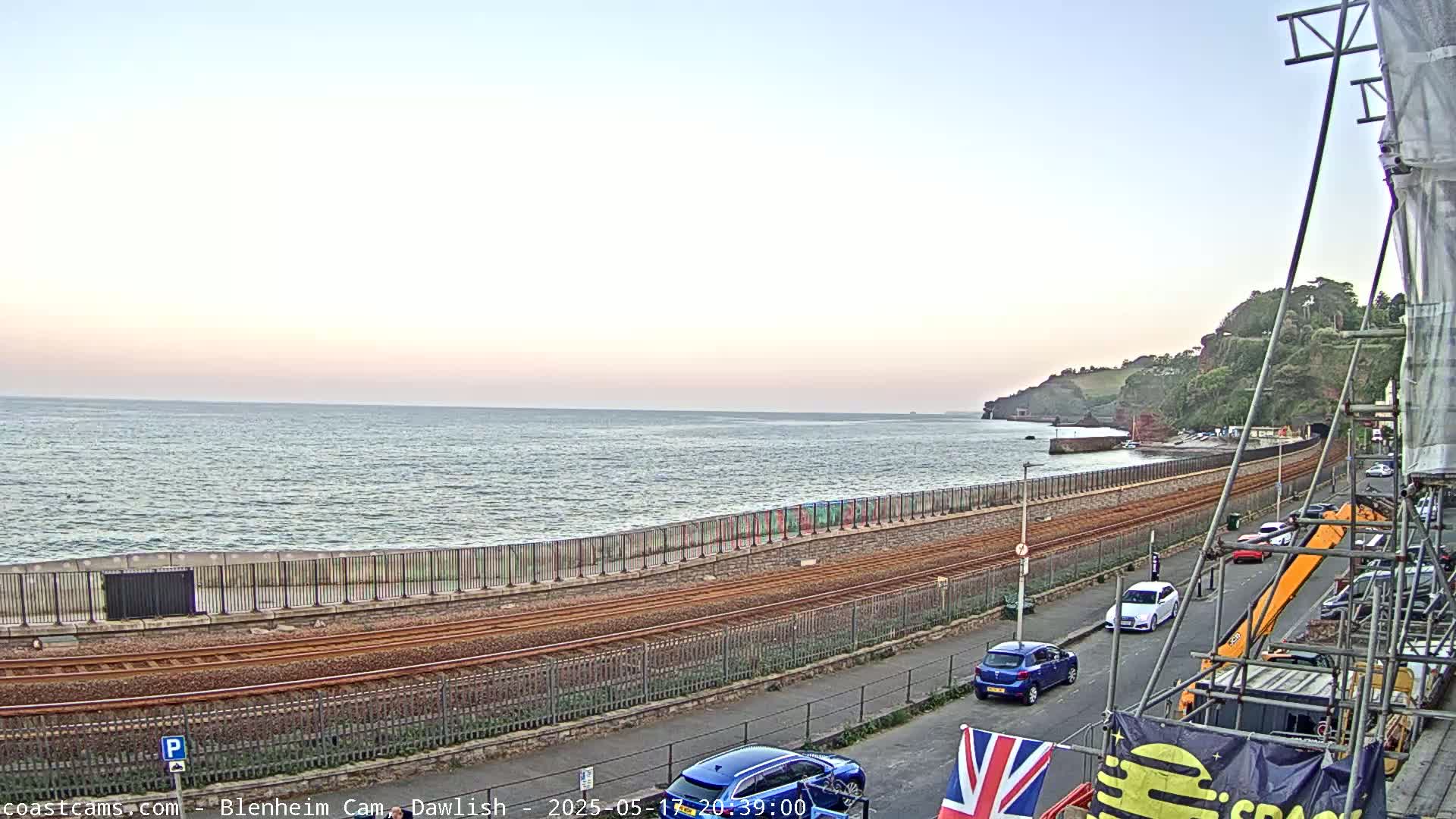 A seaside view shows calm ocean waters under a pale sky, with a railway track running alongside a road, past parked cars and construction equipment, towards a cliff.