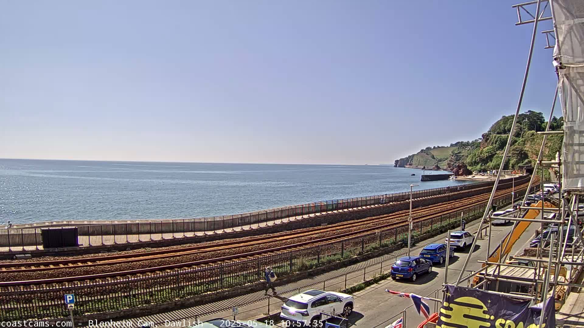 A coastal railway line runs alongside a calm ocean under a clear blue sky, with cars parked along a road next to it and a construction site visible in the foreground.