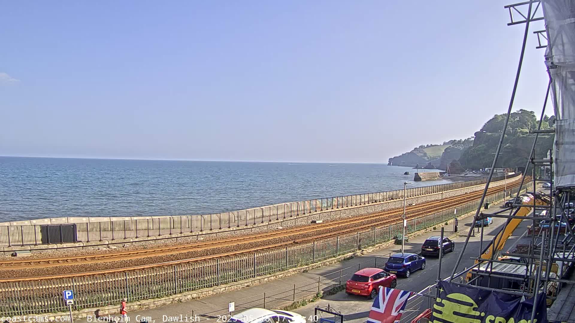 A coastal railway line runs alongside a sea wall, with cars parked nearby, under a clear blue sky.