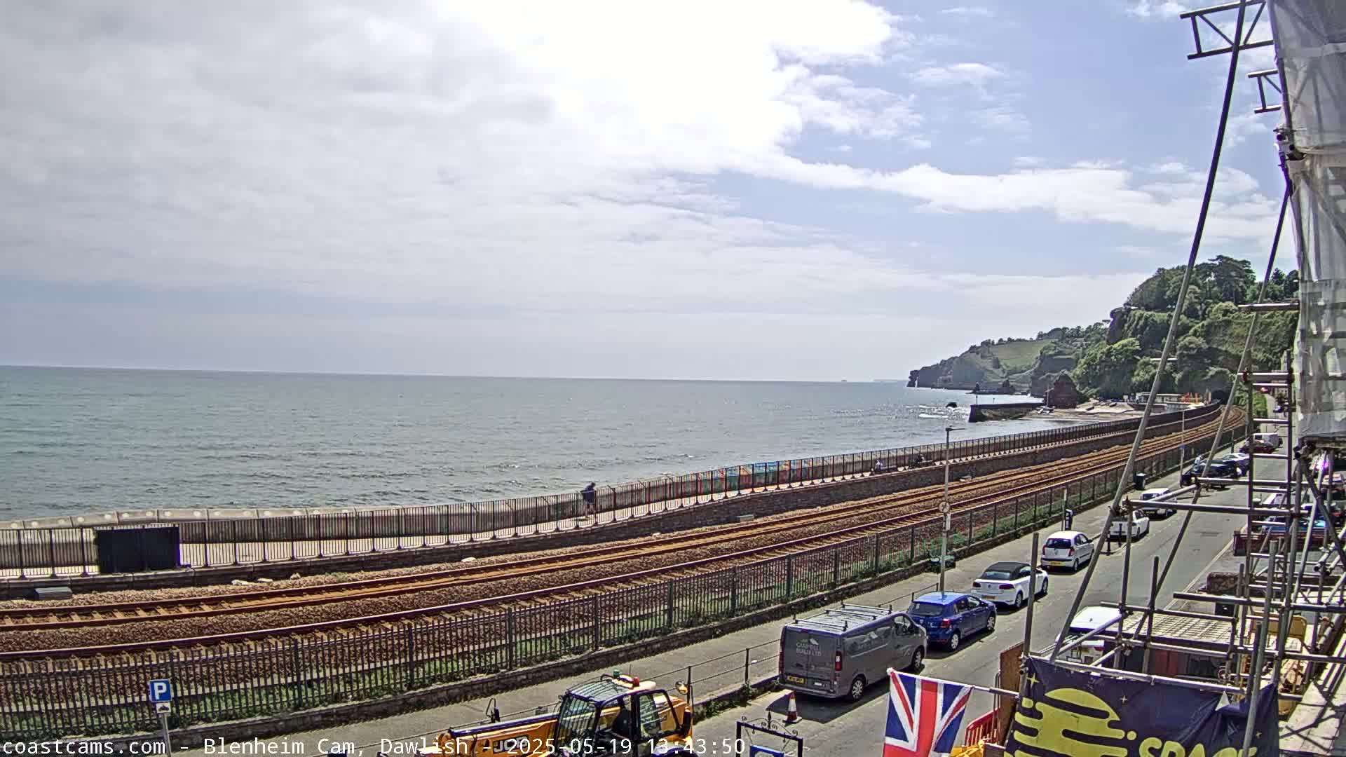 A partly cloudy day shows a coastal railway line next to a sea wall, with a beach and cliffs in the background, and several vehicles parked along the adjacent road.