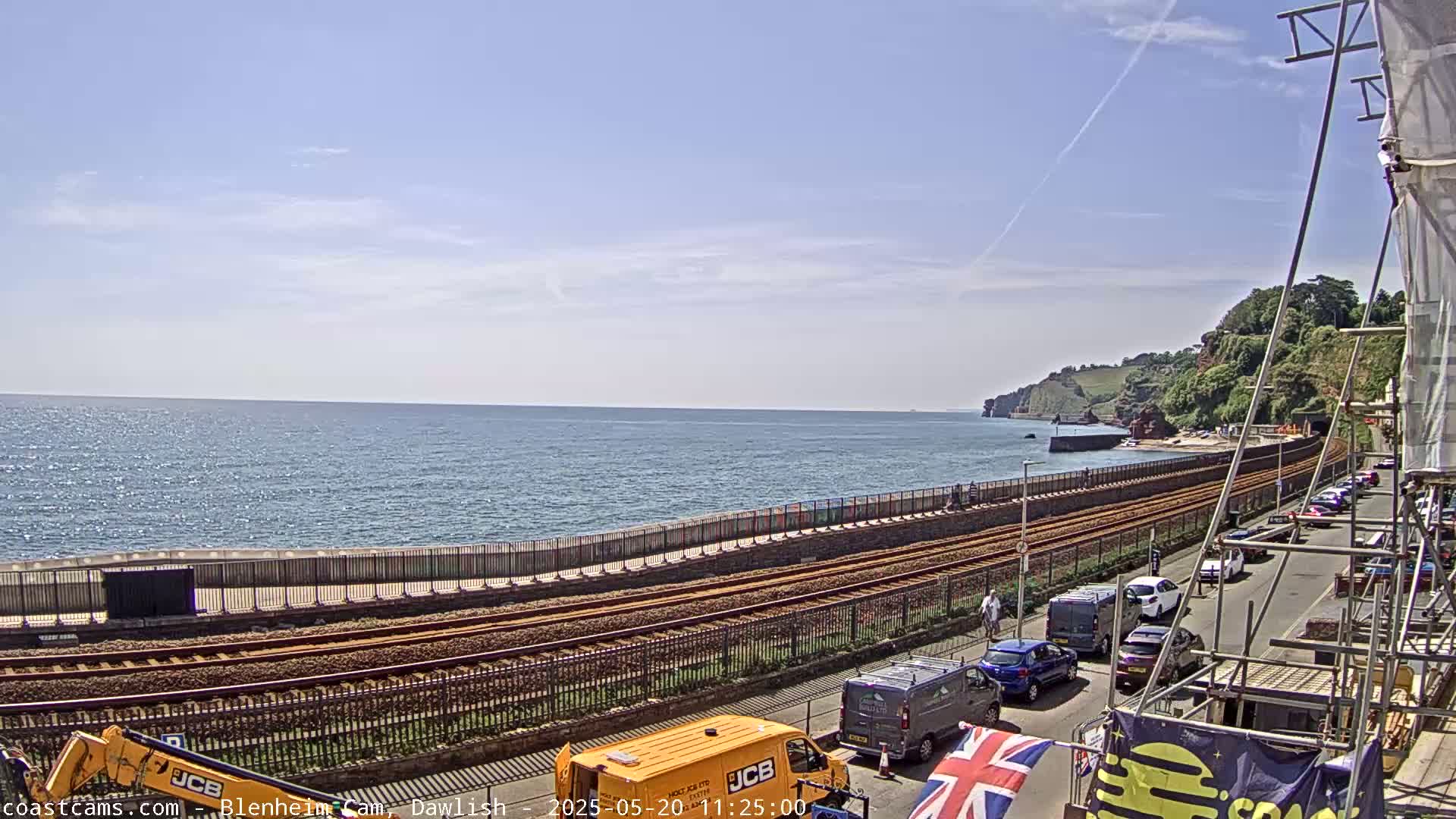 A seaside scene under a mostly clear sky shows train tracks running along a coastal path next to a calm ocean, with cars parked along a road beside the tracks and construction equipment present.