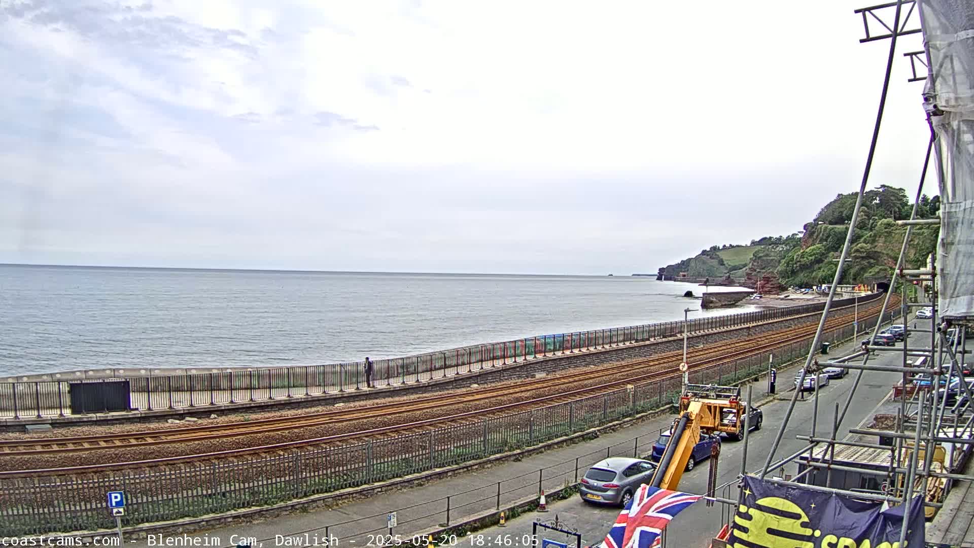 A coastal railway line runs alongside a sea wall, with cars parked on a street next to scaffolding and a partly cloudy sky above a calm sea.