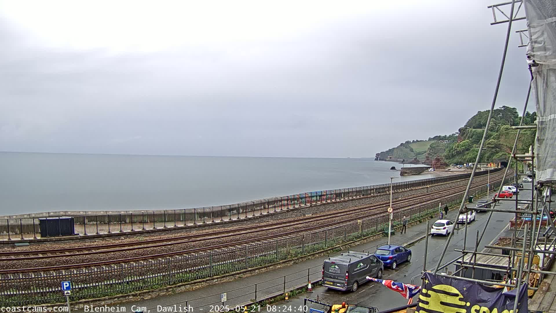 A coastal scene on an overcast day shows train tracks running alongside a sea wall bordering a calm ocean, with a road and parked cars visible beyond the tracks.