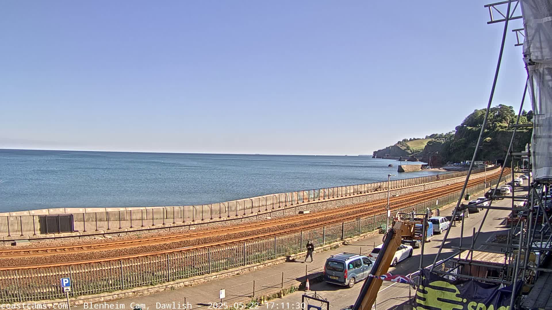 A coastal railway line runs alongside a calm sea under a clear blue sky, with cars parked along a road next to it and a few people walking nearby.