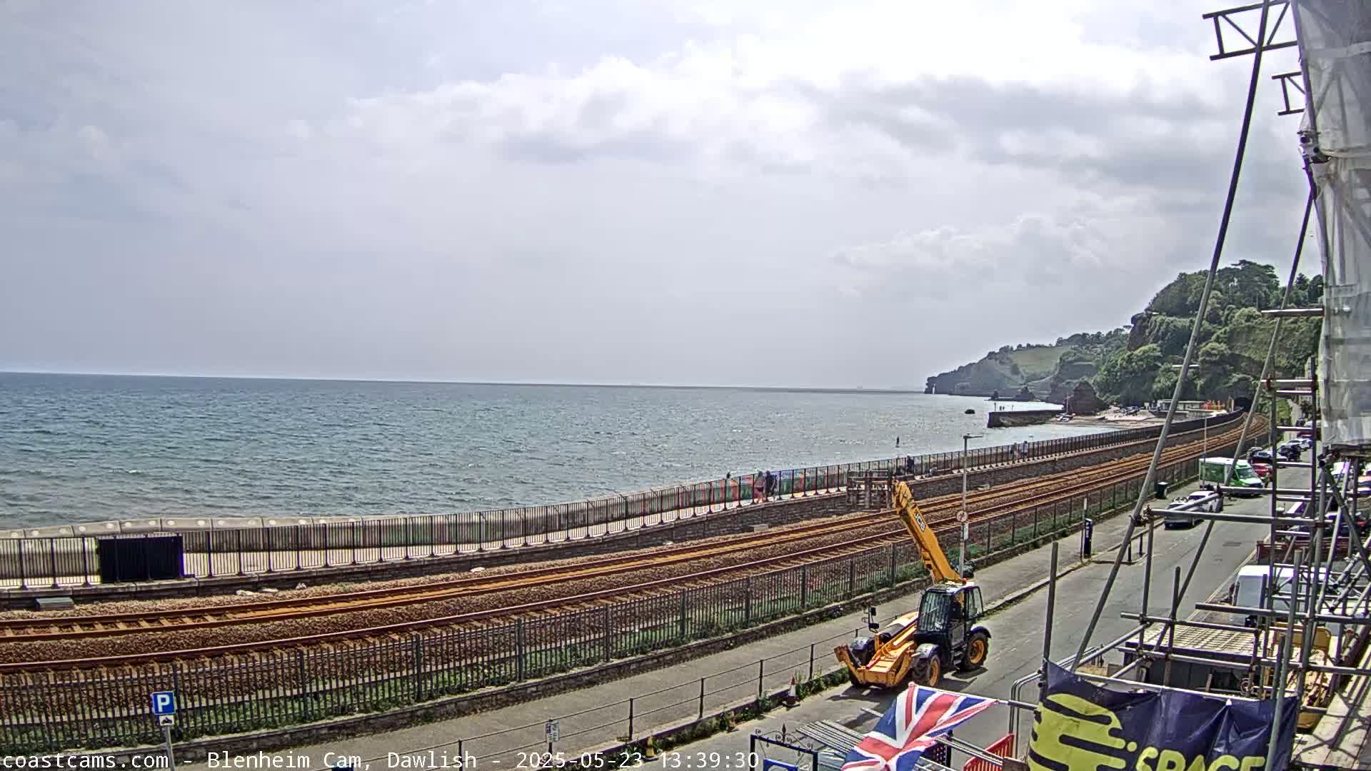 A partly cloudy day shows a seaside view of train tracks running alongside a sea wall, with a yellow telehandler parked nearby and construction scaffolding visible.
