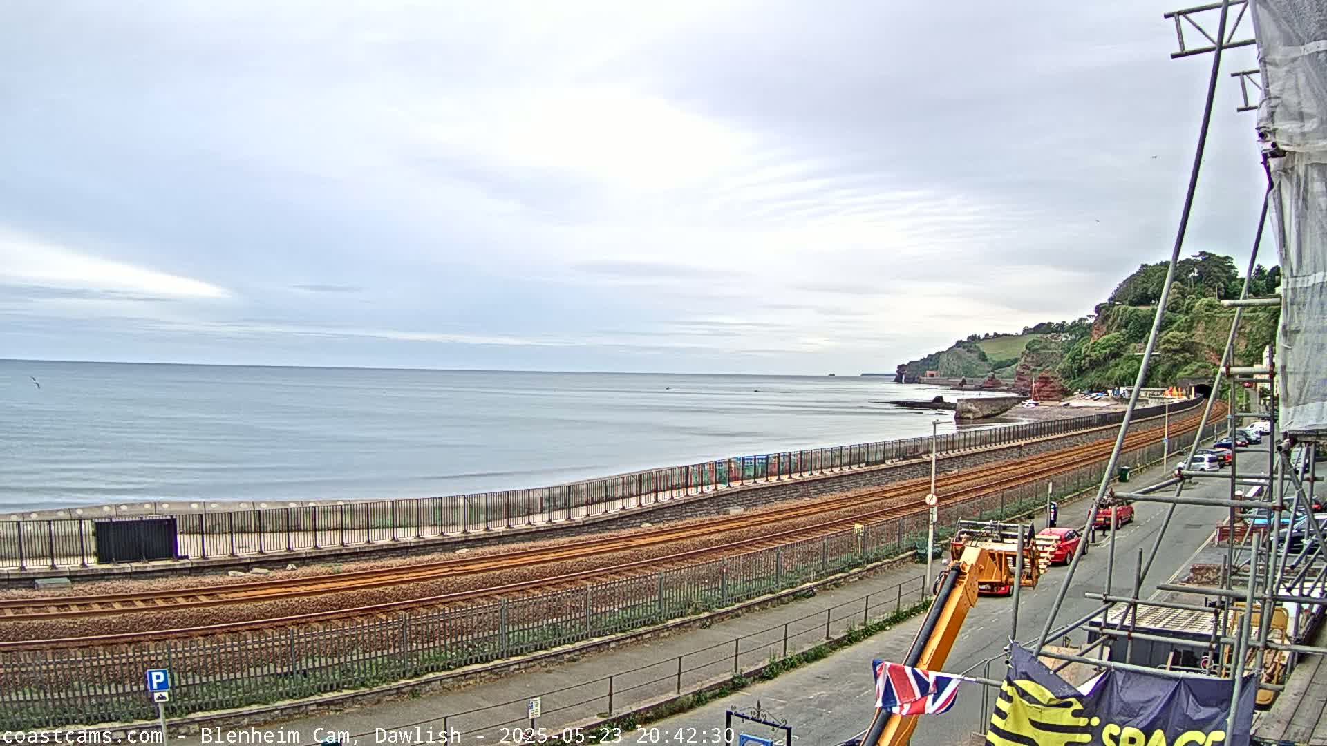A calm ocean with a cloudy sky is seen beyond a railway line that runs alongside a paved road and a seawall, with some cars parked along the road and construction equipment present.