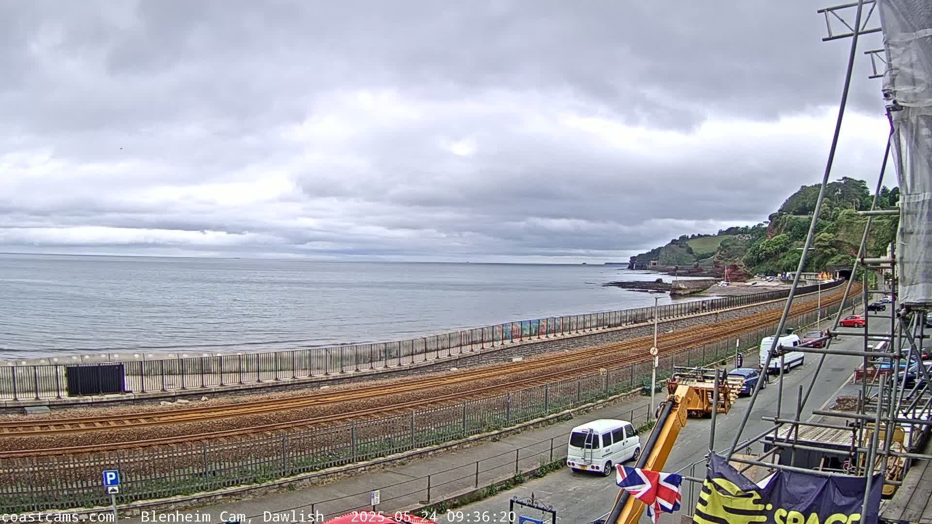 A coastal scene under an overcast sky shows train tracks running alongside a beach, with a road and construction scaffolding visible in the foreground.