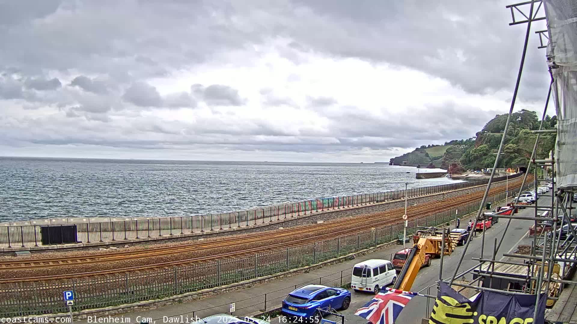 A coastal scene under an overcast sky shows a train track running alongside a seawall, with several cars parked along a street next to it, and a body of water stretching to the horizon.