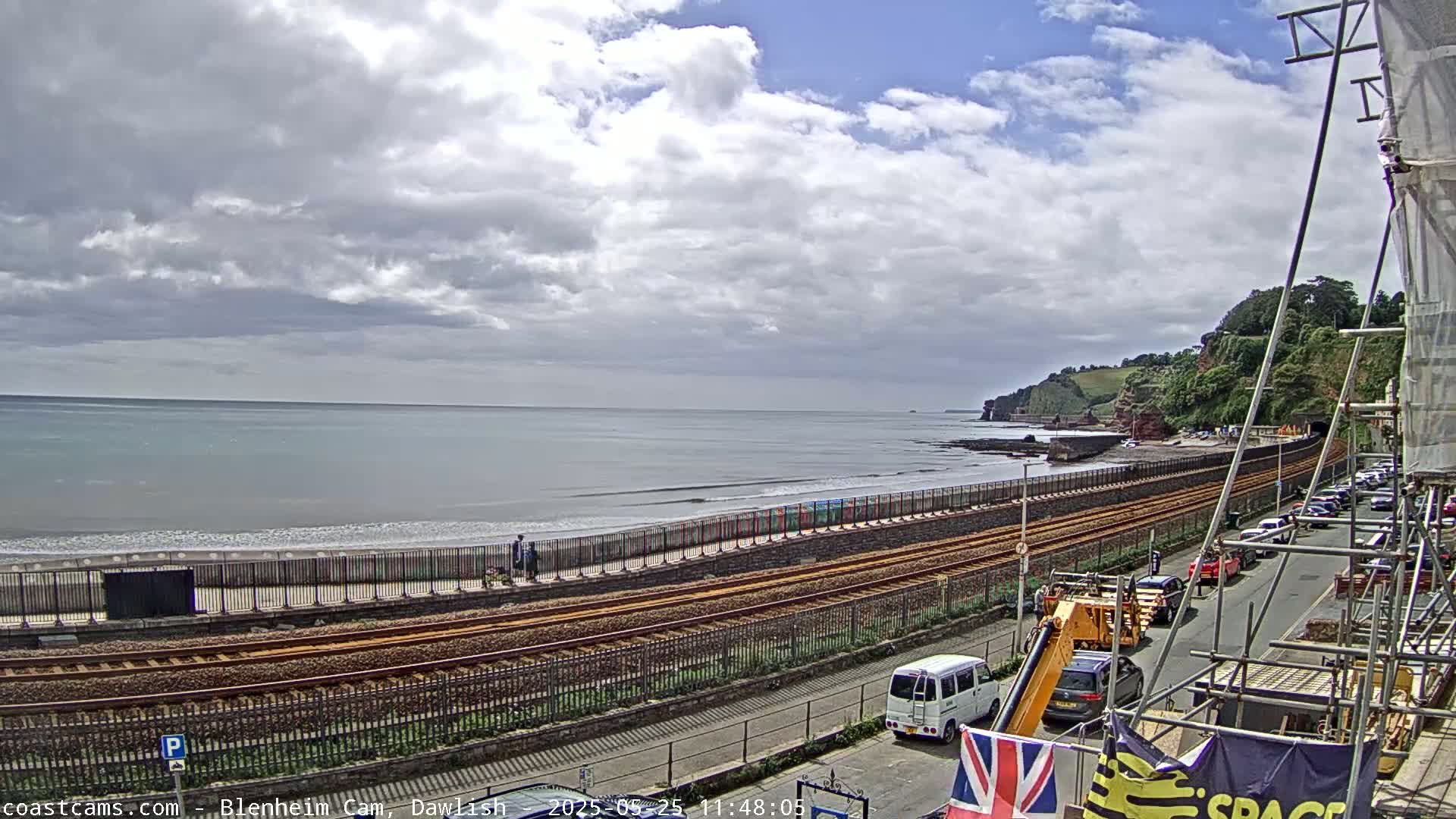 A partly cloudy day shows a beach with calm waves, a railway line running alongside it, a road with parked cars, and green hills in the background.