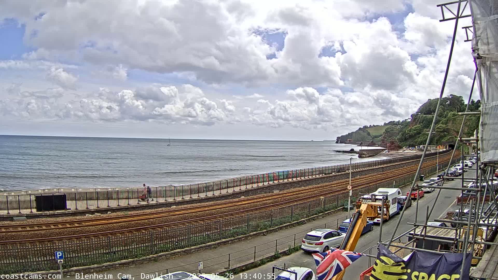 A partly cloudy day overlooks a coastline with a railway track running alongside a road, and parked cars visible in front of buildings under construction.