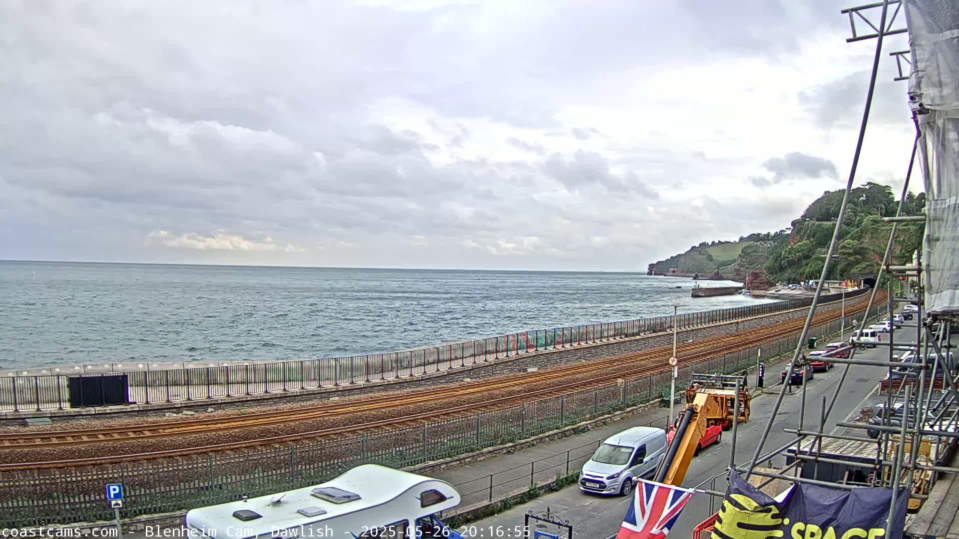 A coastal scene under an overcast sky shows a railway line running alongside a seawall, with a road and parked vehicles beyond, and a body of water with small waves in the background.