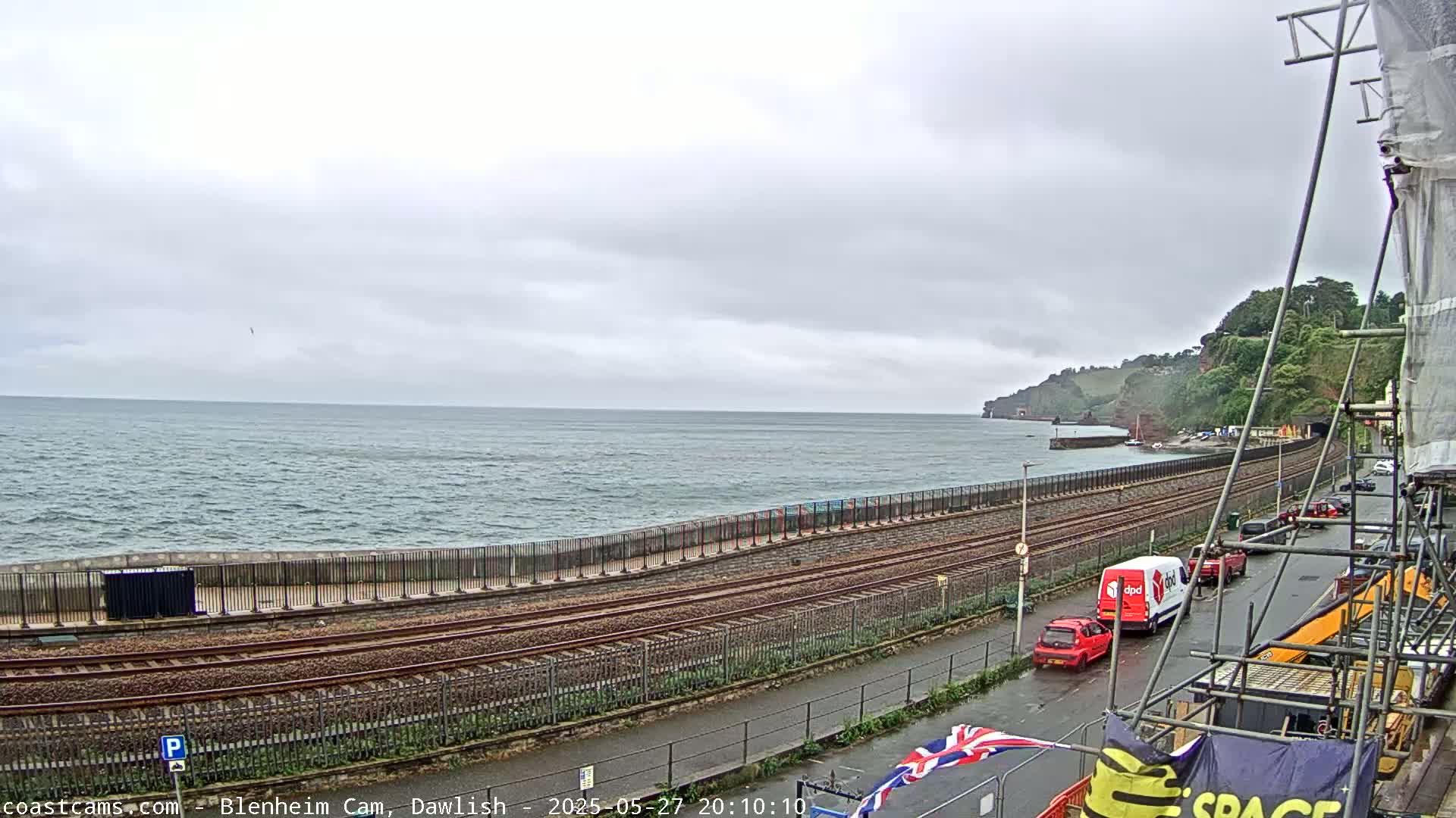 A coastal scene under an overcast sky shows a railway track running alongside a sea wall, with a few vehicles parked on a road next to it, and a rocky cliffside in the distance.