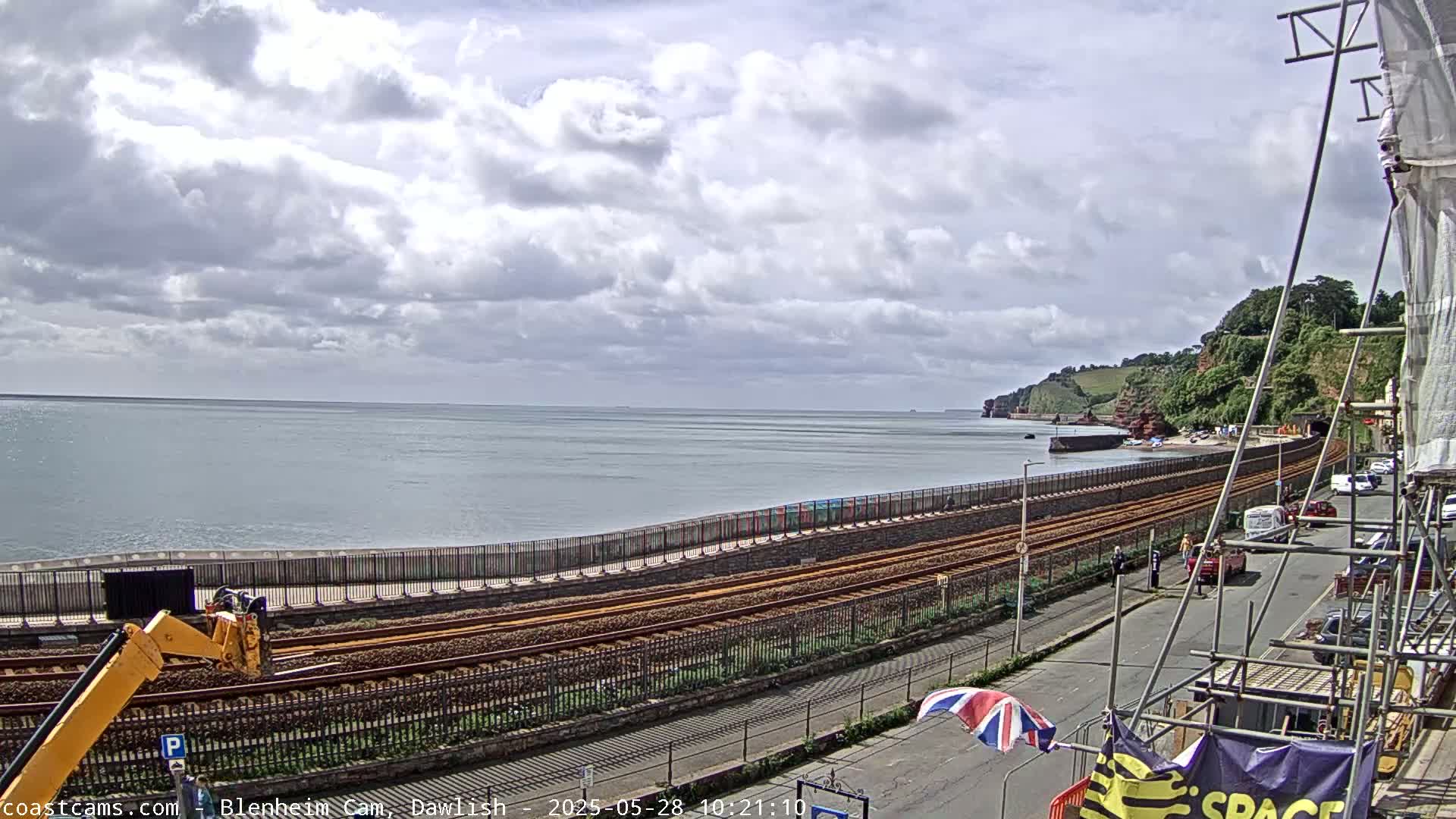 A partly cloudy day overlooks a coastal railway line next to a beach and calm ocean, with construction scaffolding visible in the foreground.