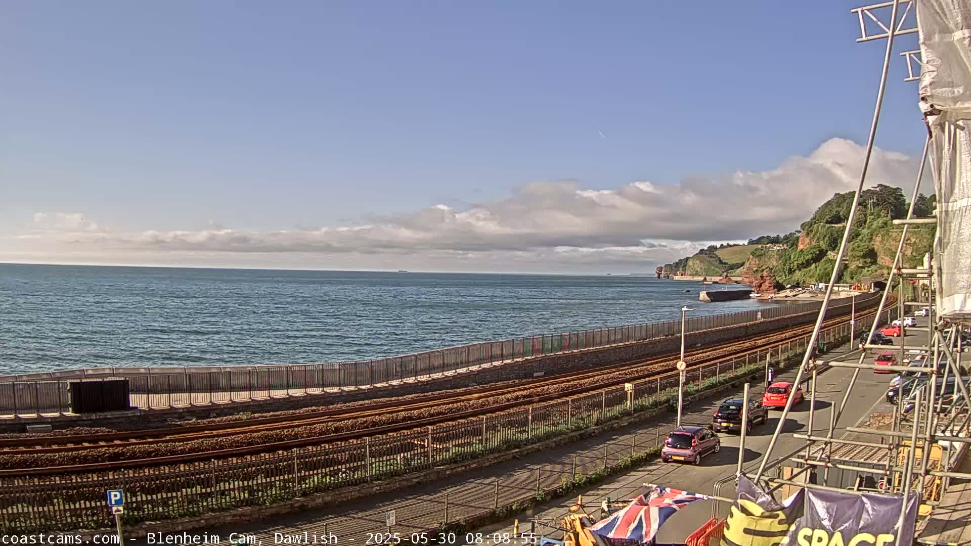 A partly cloudy day overlooks a coastline with a railway track running alongside a road where several cars are parked,  all situated next to a body of water.