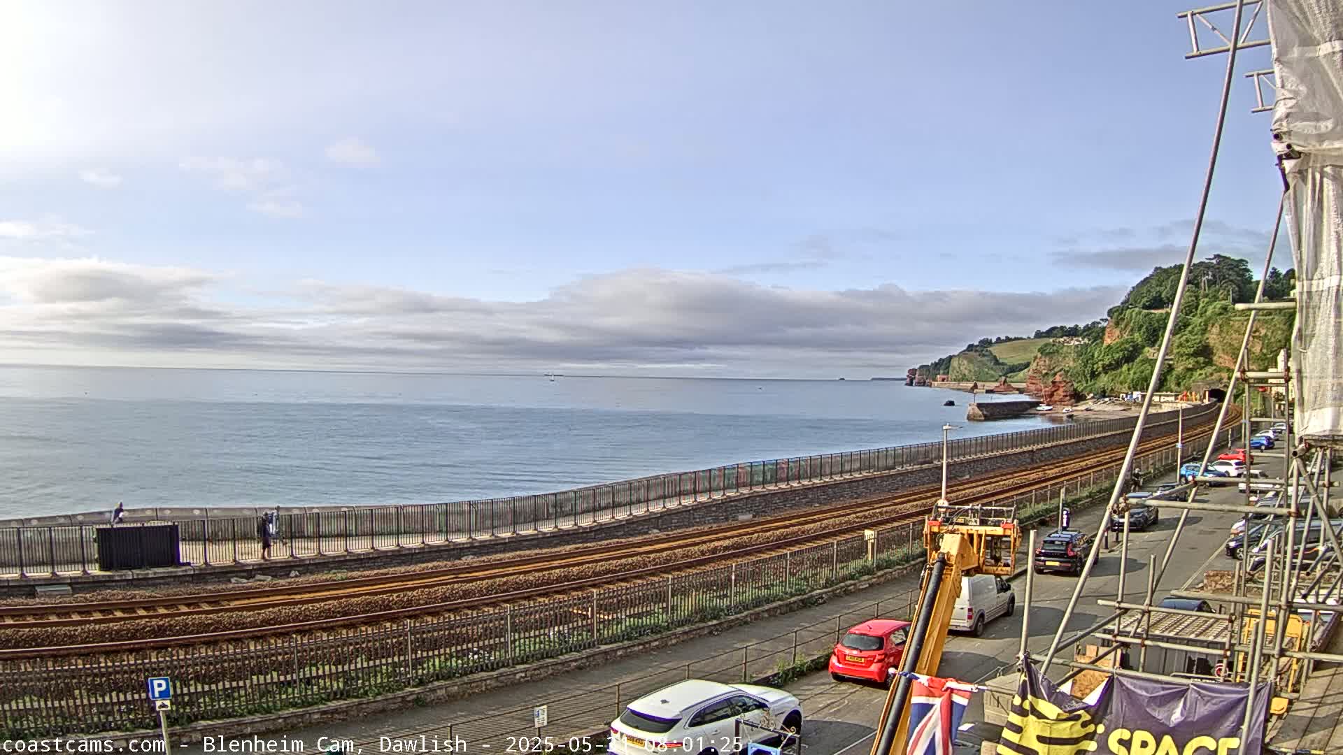 A partly cloudy day reveals a coastal scene with a railway line running alongside a seawall, overlooking a calm ocean and a line of parked cars on a street next to the tracks.