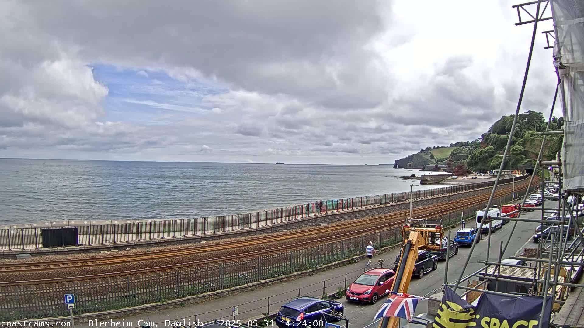 A mostly cloudy day overlooks a seaside town with a railway track running along the coastline, where cars are parked along a street next to construction scaffolding.