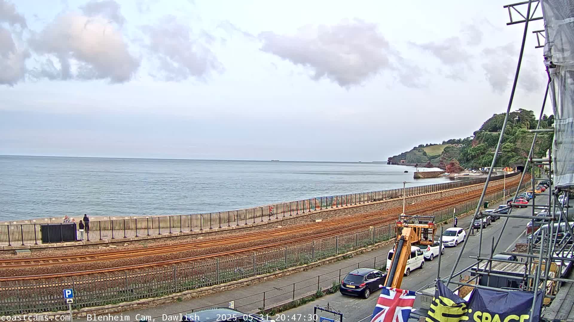 A seaside view shows train tracks running alongside a seawall, with cars parked on a street above and a partly cloudy sky.