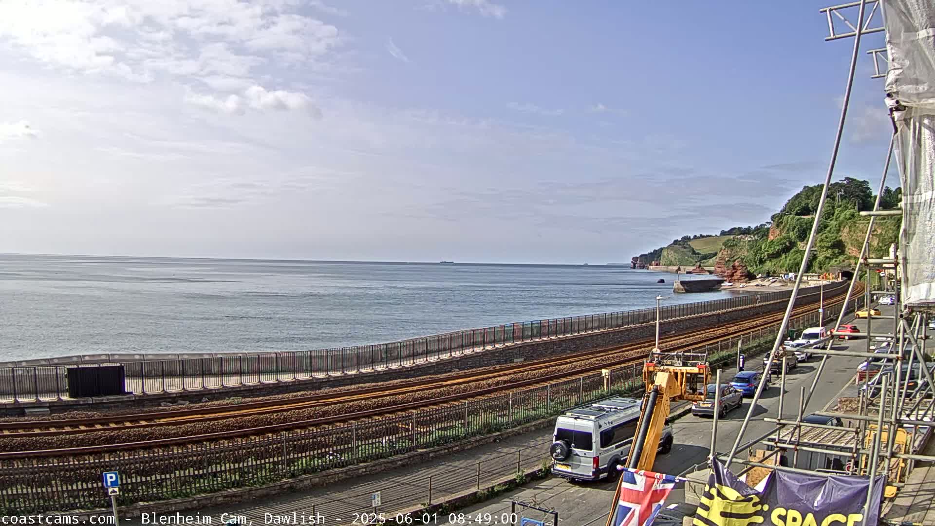 A partly cloudy day shows a seaside town with train tracks running alongside a seawall, a road with parked cars, and construction scaffolding.