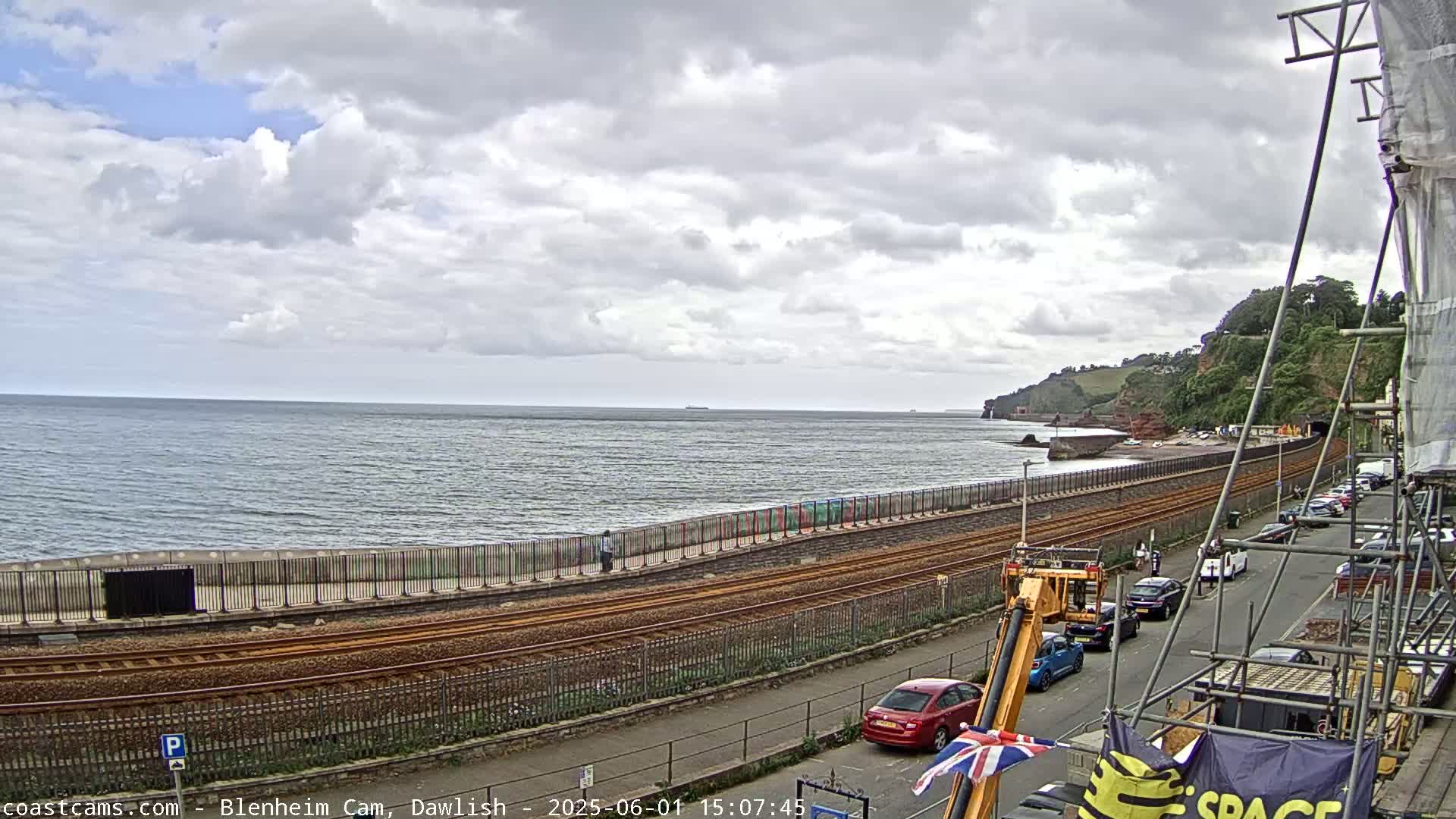 A coastal railway line runs alongside a sea wall, with a partly cloudy sky overhead and several cars parked on a street next to the tracks.