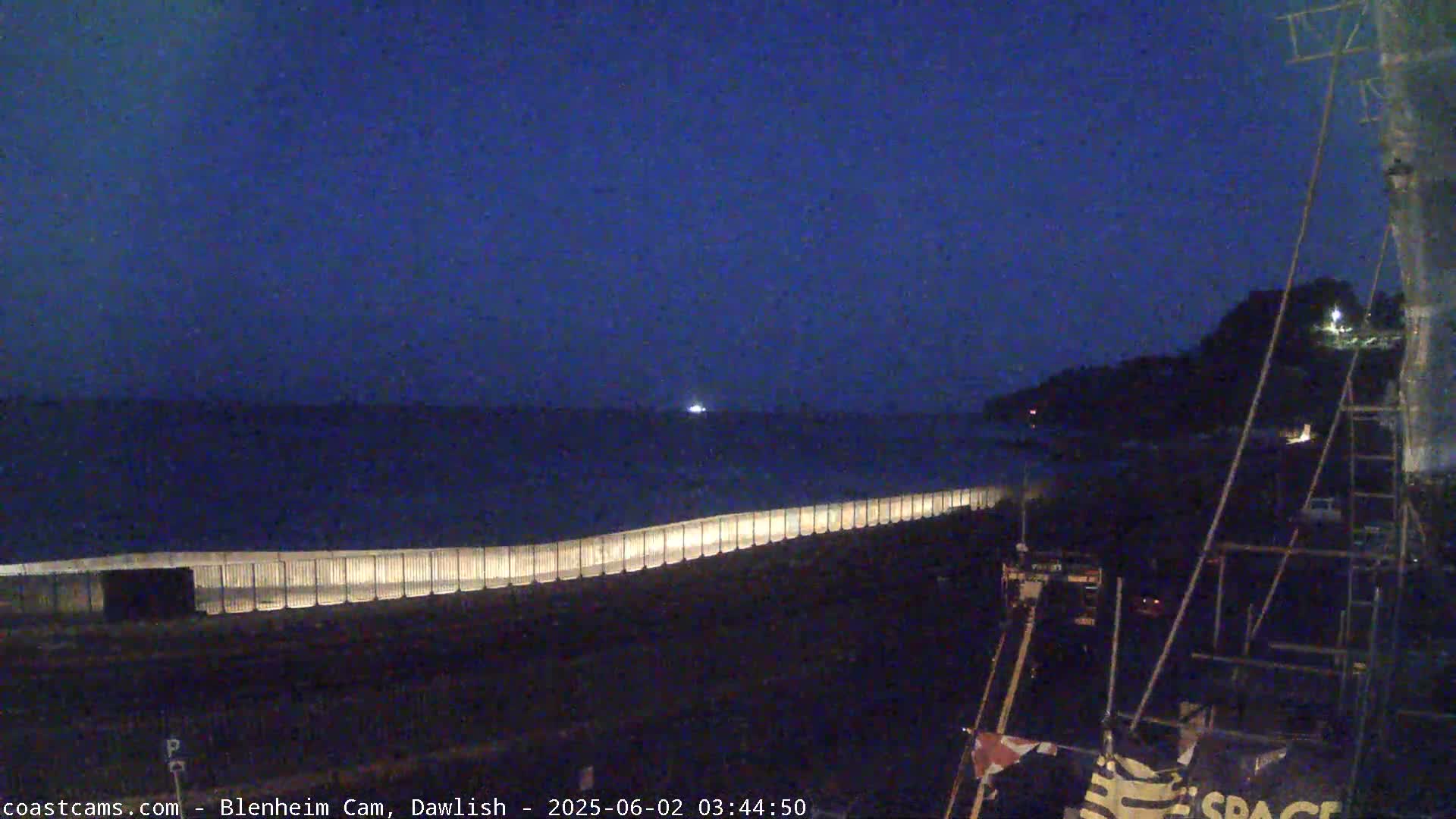A dark, nighttime scene shows a lit seawall extending into a calm, dark ocean under a clear, deep blue sky.
