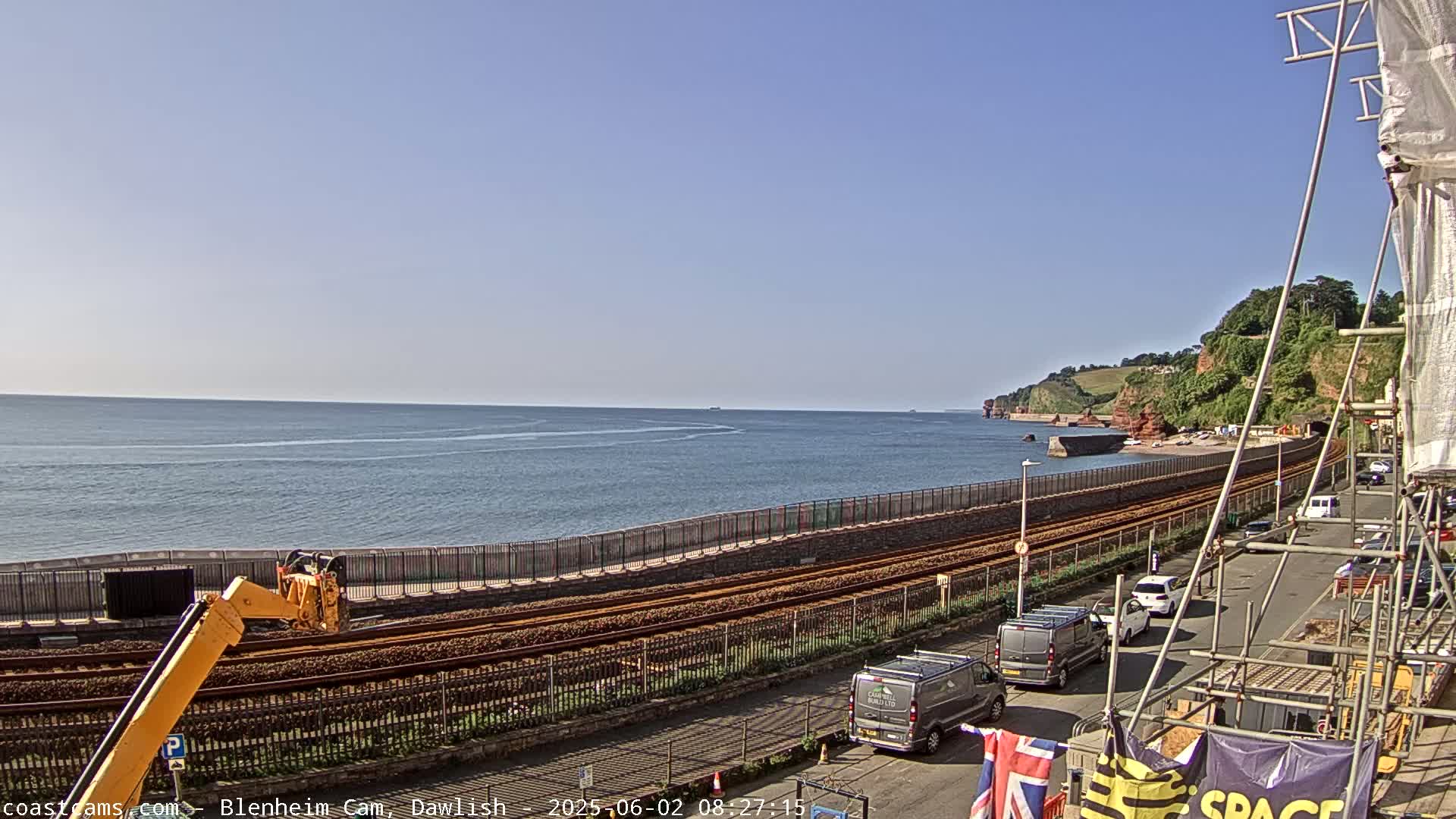 A clear day reveals a coastal scene with a railway line running alongside a sea wall, construction equipment present, and several vehicles parked along a road overlooking the ocean.