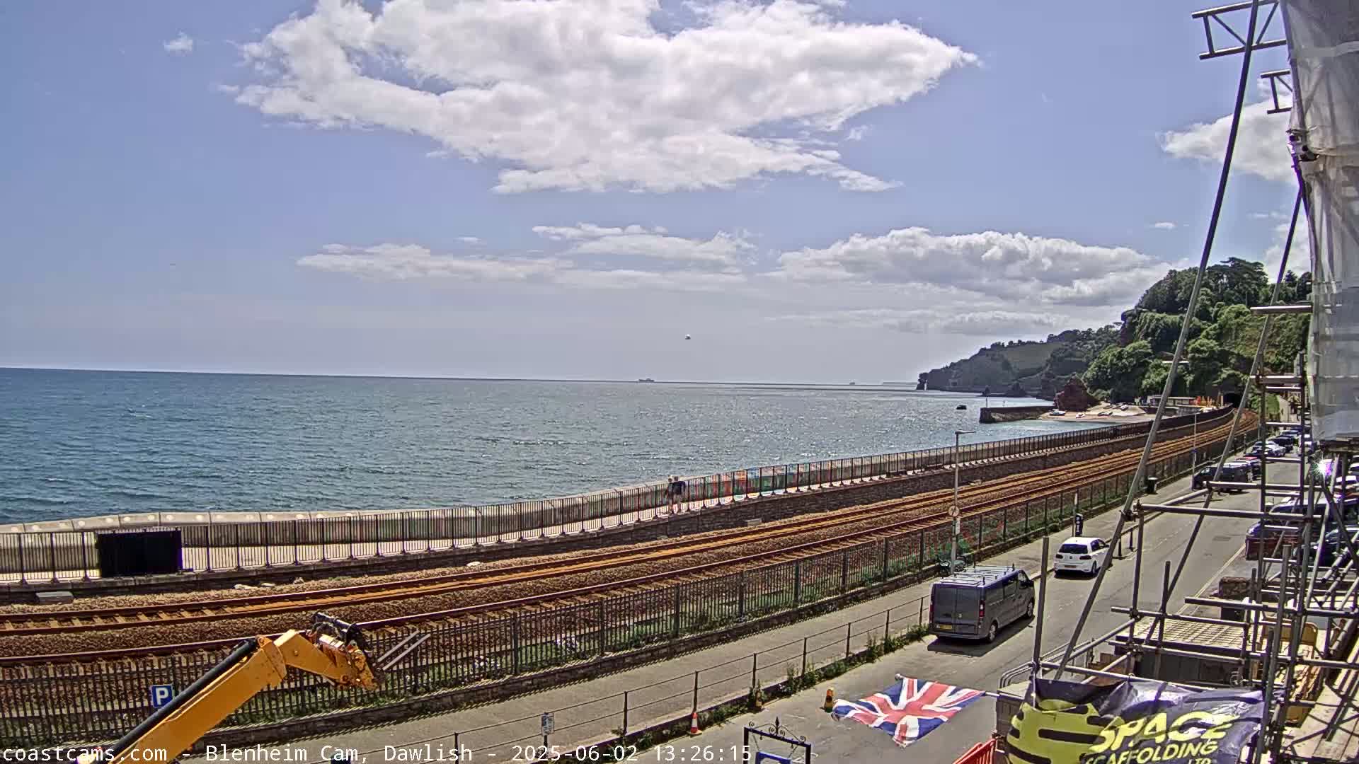 A partly cloudy day shows a coastal railway track running alongside a seawall, with vehicles parked along a road next to it, and the ocean in the background.