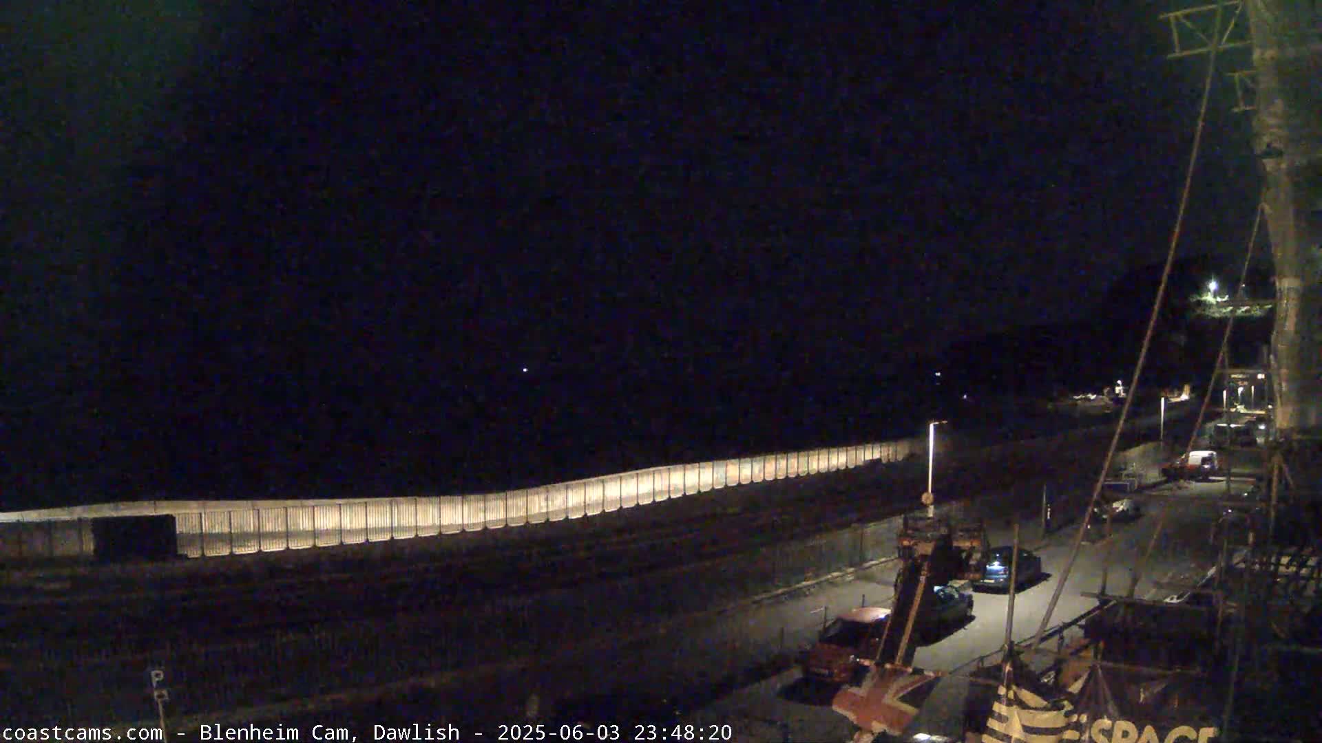 A nighttime, long shot of a seawall illuminated with lights, a few vehicles in a parking lot, and a dark sky.