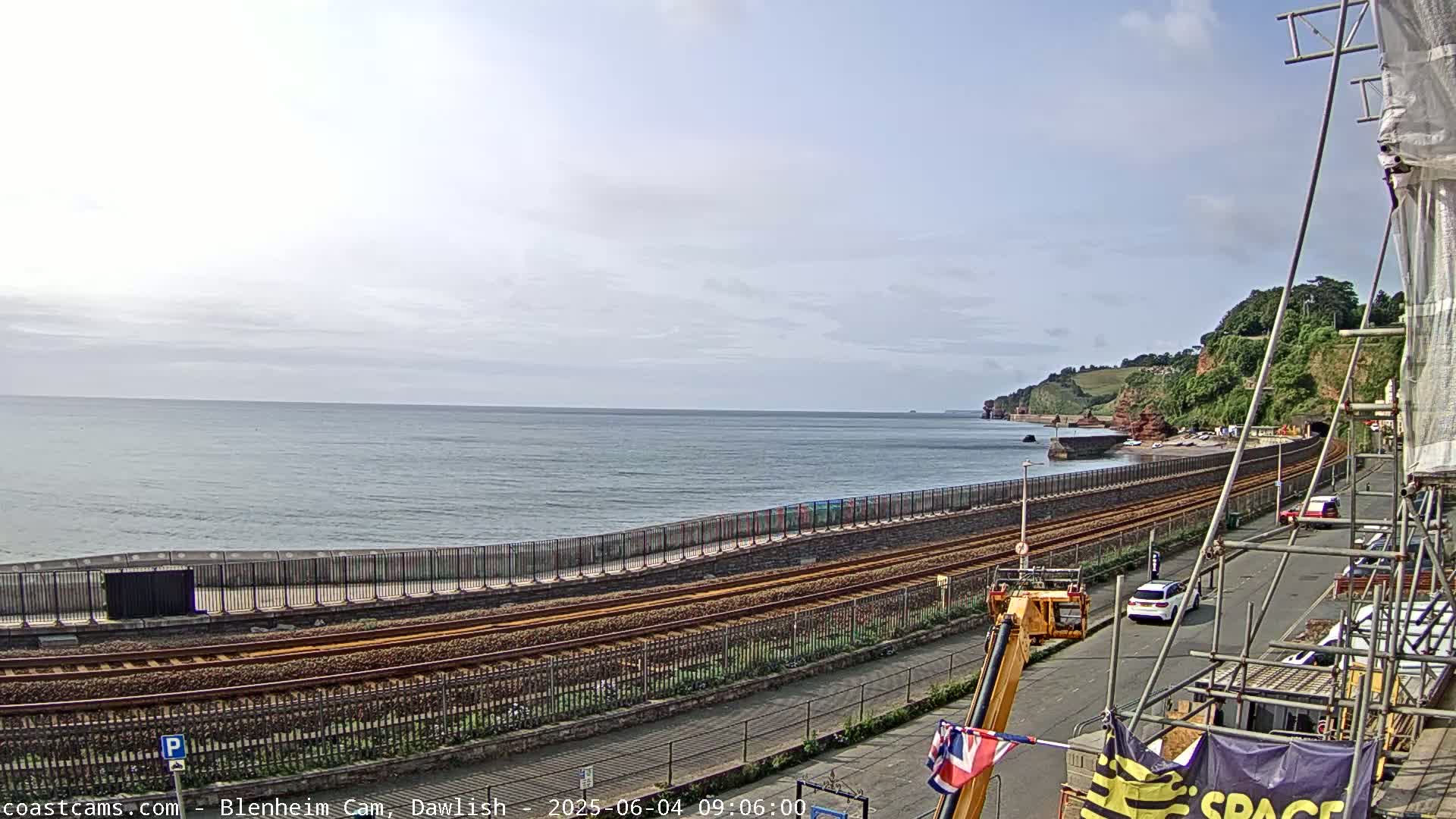 A coastal railway line runs alongside a sea wall, with a calm sea and a partly cloudy sky visible in the background.