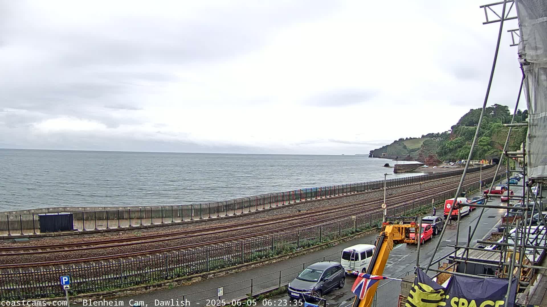 A seaside railway line runs alongside a gray, overcast ocean, bordered by a road with parked cars and construction scaffolding.