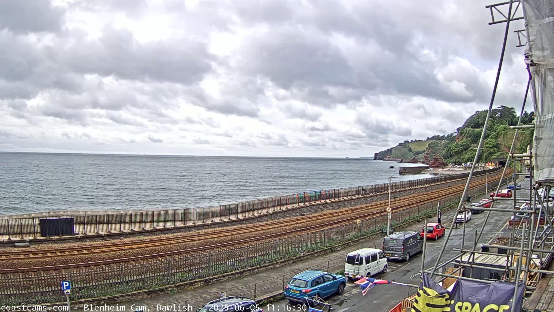 A coastal railway line runs alongside a sea wall, with parked cars lining a street on the other side, under a cloudy sky.