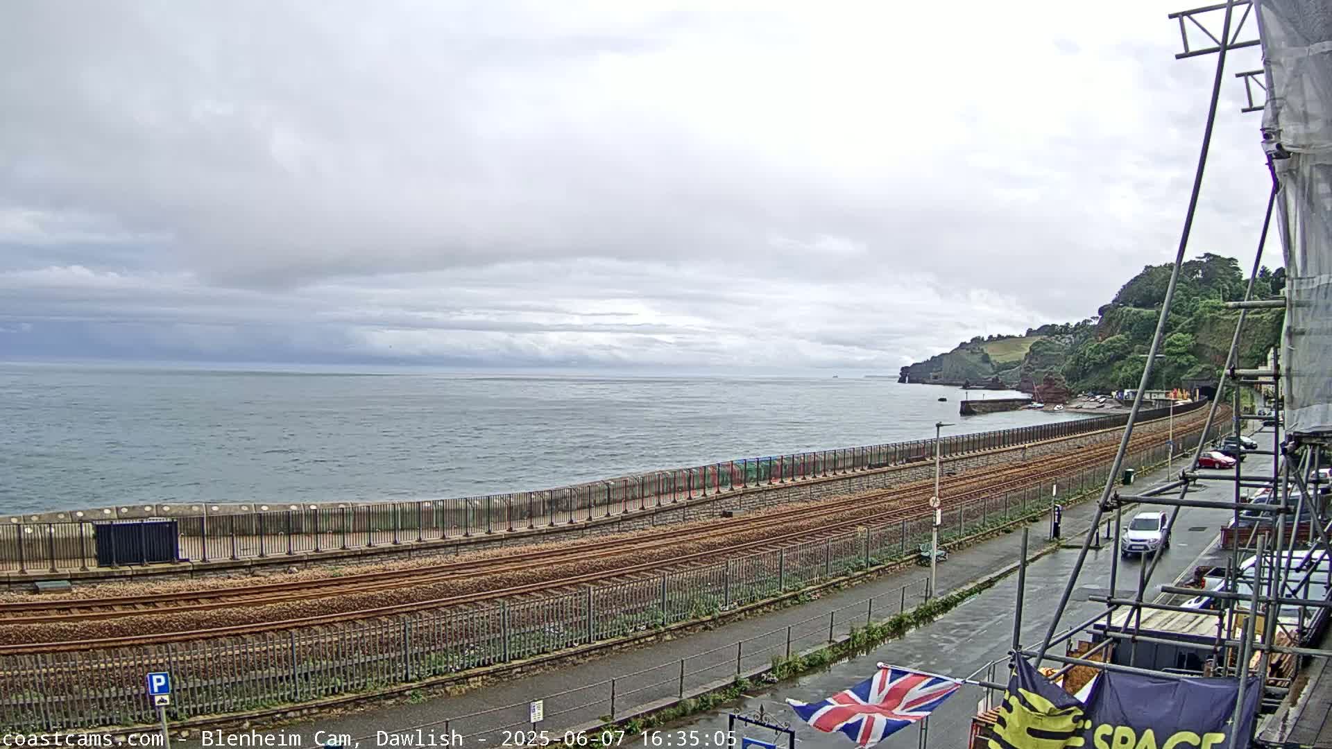 A coastal railway line runs alongside a sea wall, with a calm sea and overcast sky in the background, and a street with cars parked along the side.