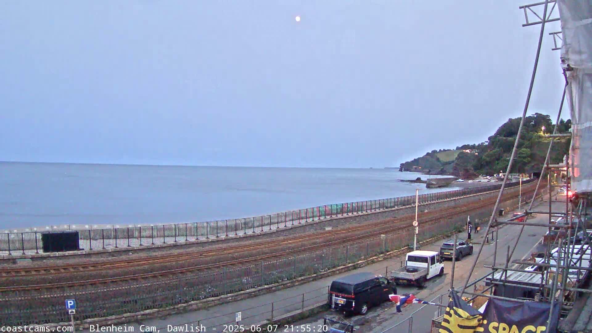 A calm sea stretches out beneath a pale, moonlit sky, next to a coastal railway line and road with a few cars parked alongside a seawall, backed by green hills.