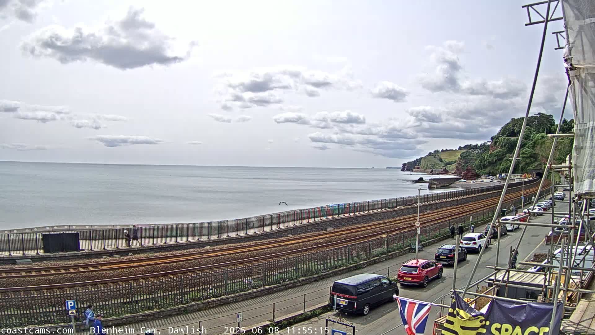 A coastal scene under a partly cloudy sky shows a railway line running parallel to a seawall and road, with parked cars and some pedestrians visible near the water.