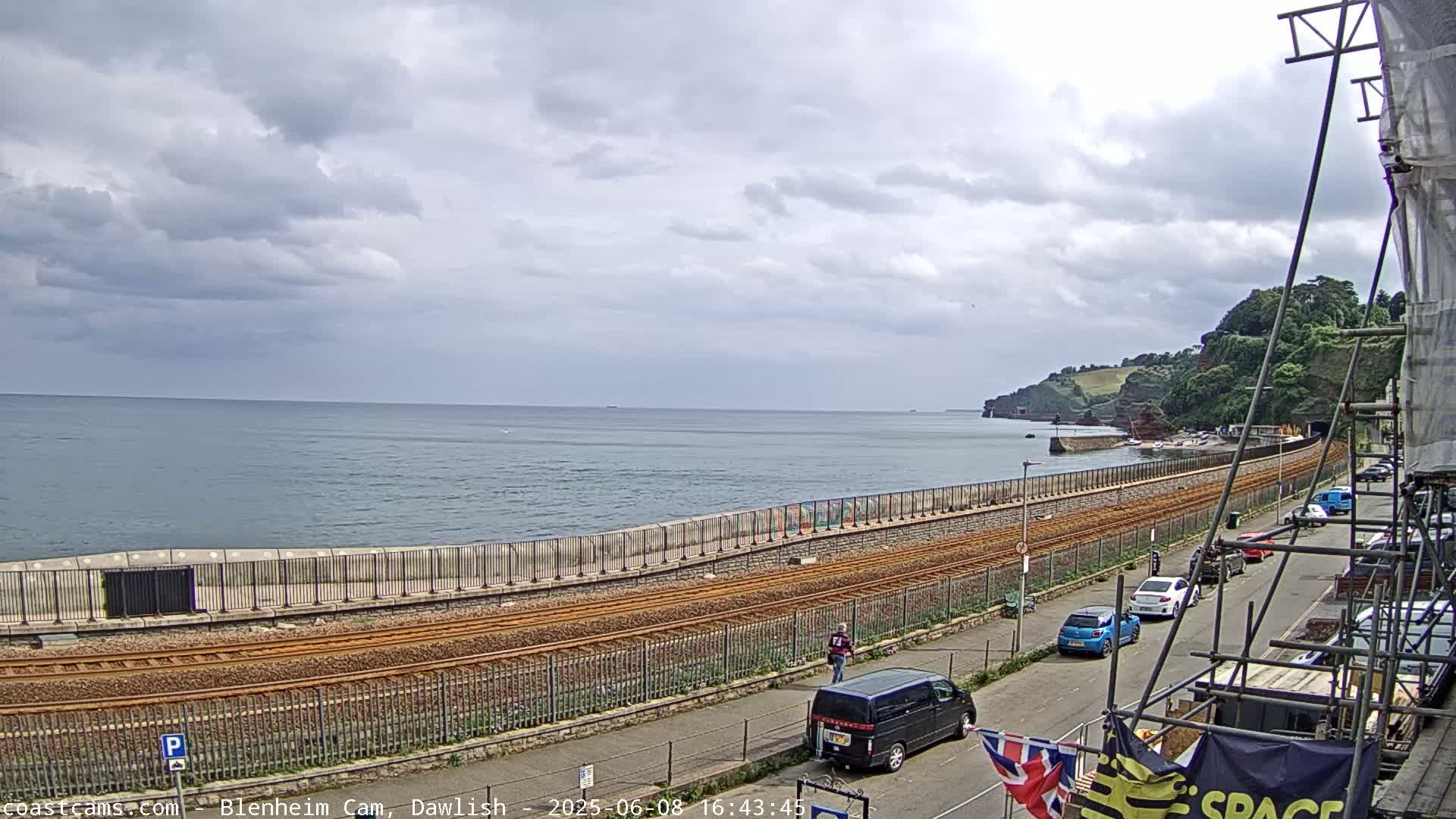 A coastal scene under a cloudy sky shows train tracks running alongside a sea wall, with cars parked on a street and a body of water extending to the horizon.