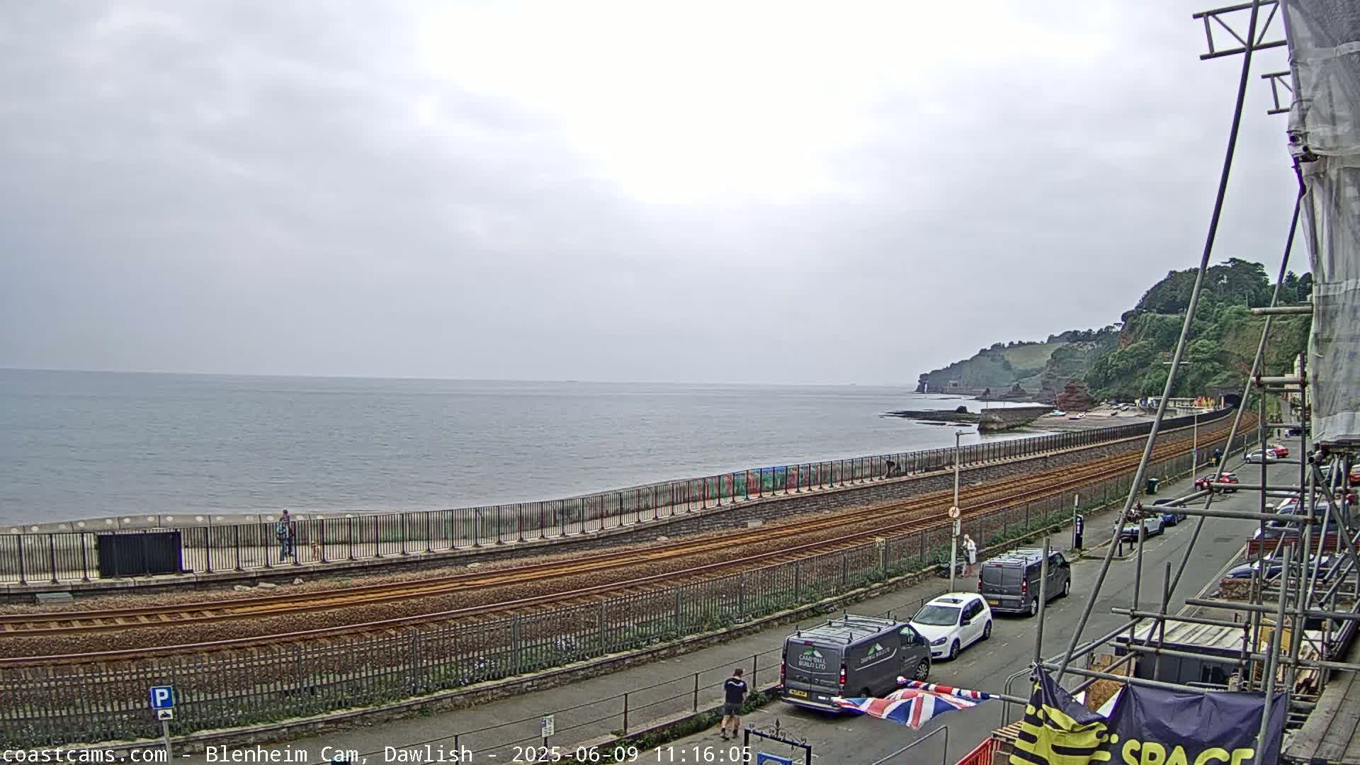 A mostly cloudy day reveals a coastal scene with train tracks running alongside a seawall, a beach, and parked cars lining a street next to construction scaffolding.