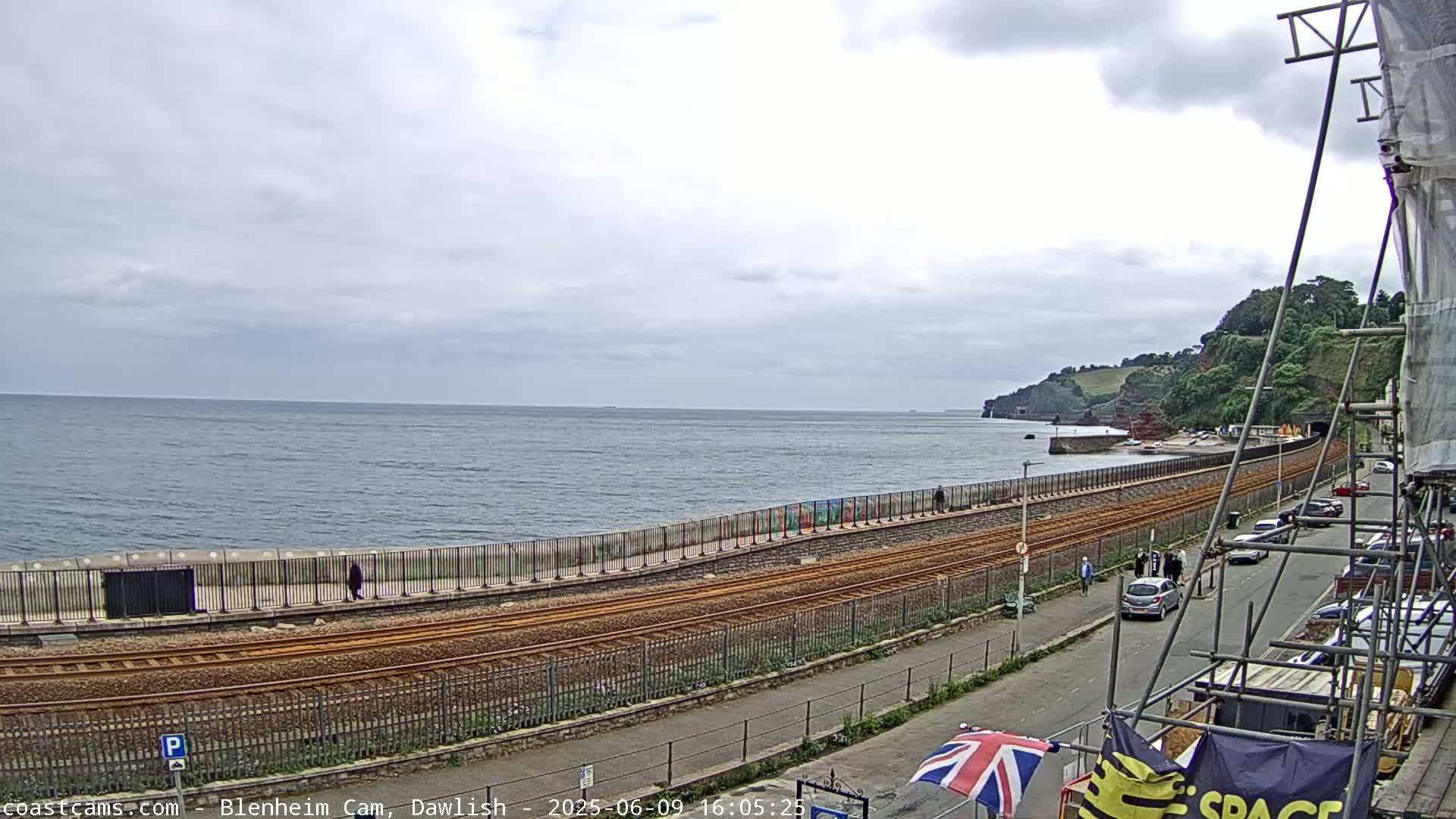 A coastal scene on an overcast day shows train tracks running alongside a seawall, with a road and parked cars beyond, and a body of water extending to cliffs in the distance.