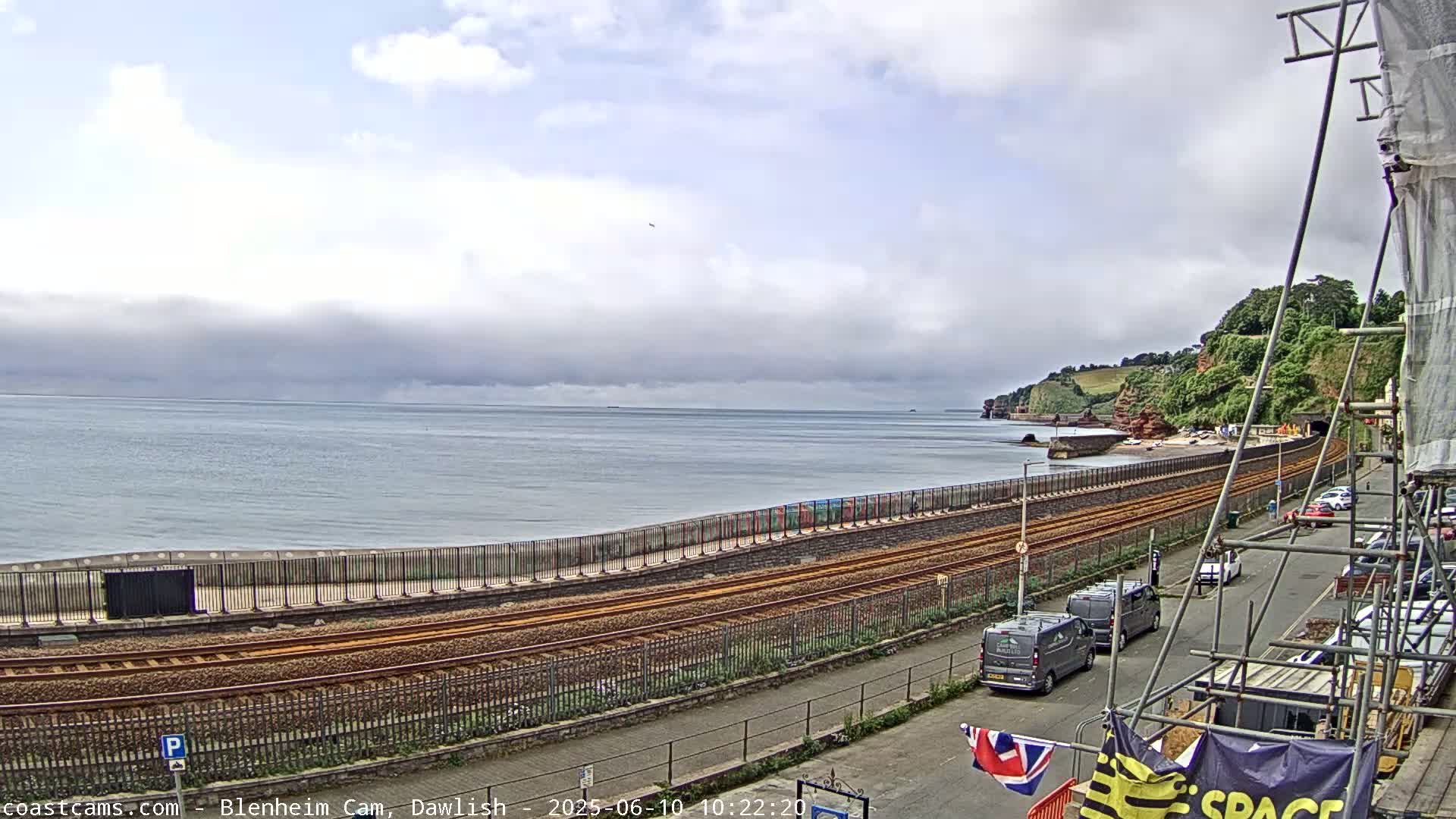 A mostly cloudy day overlooks a coastal railway line next to a beach and a seaside town, with parked cars and scaffolding visible.