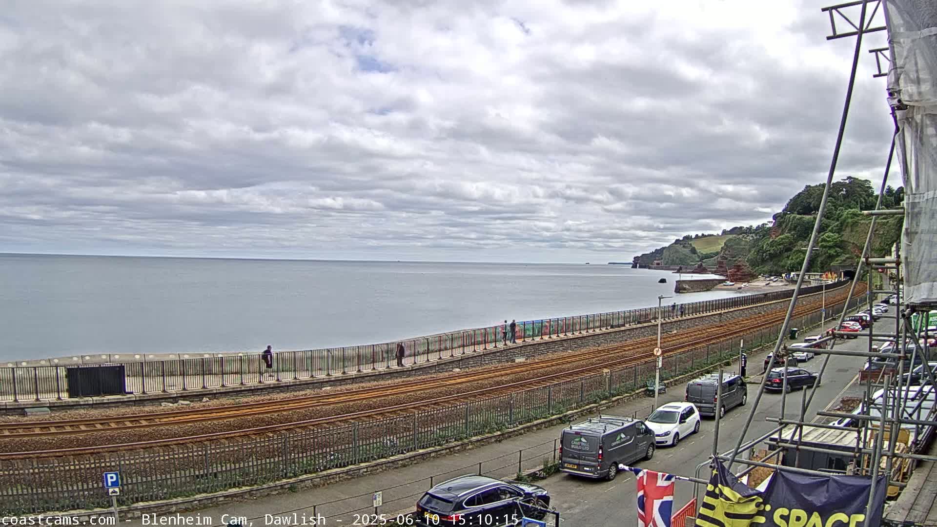 A coastal scene under a cloudy sky shows train tracks running alongside a seawall with pedestrians and parked cars nearby.