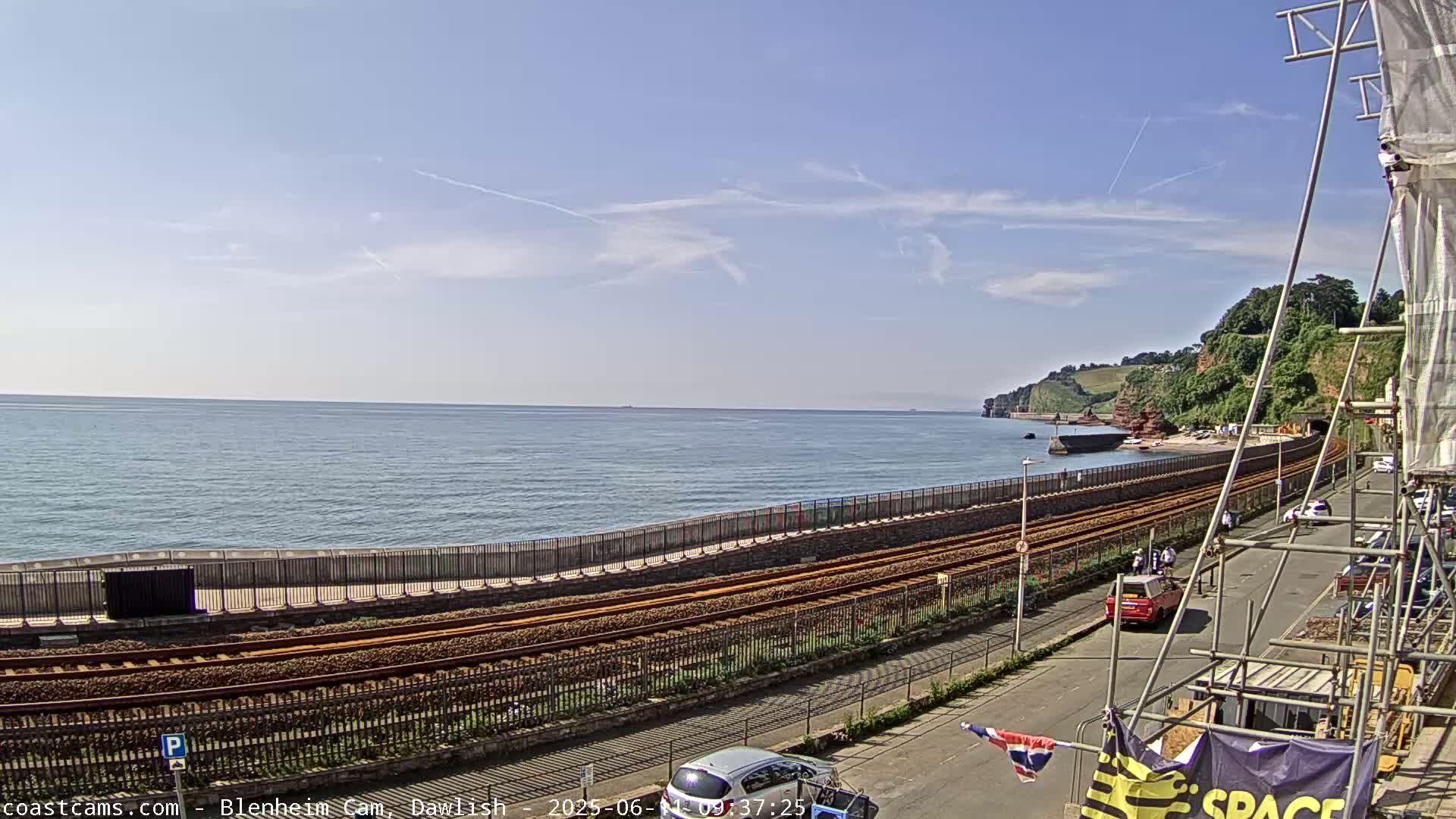 A coastal scene on a sunny day shows train tracks running alongside a seawall separating the tracks from a beach and ocean, with a partially visible town in the background.