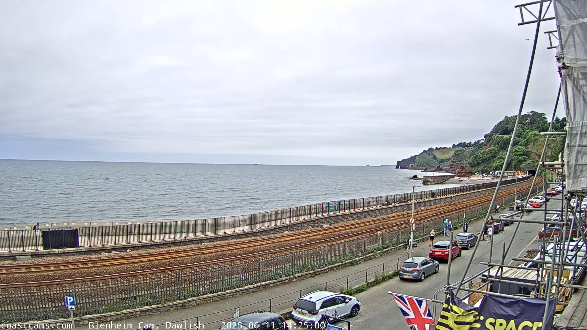 A coastal railway track runs parallel to a calm sea under an overcast sky, beside a road with parked cars and pedestrians.