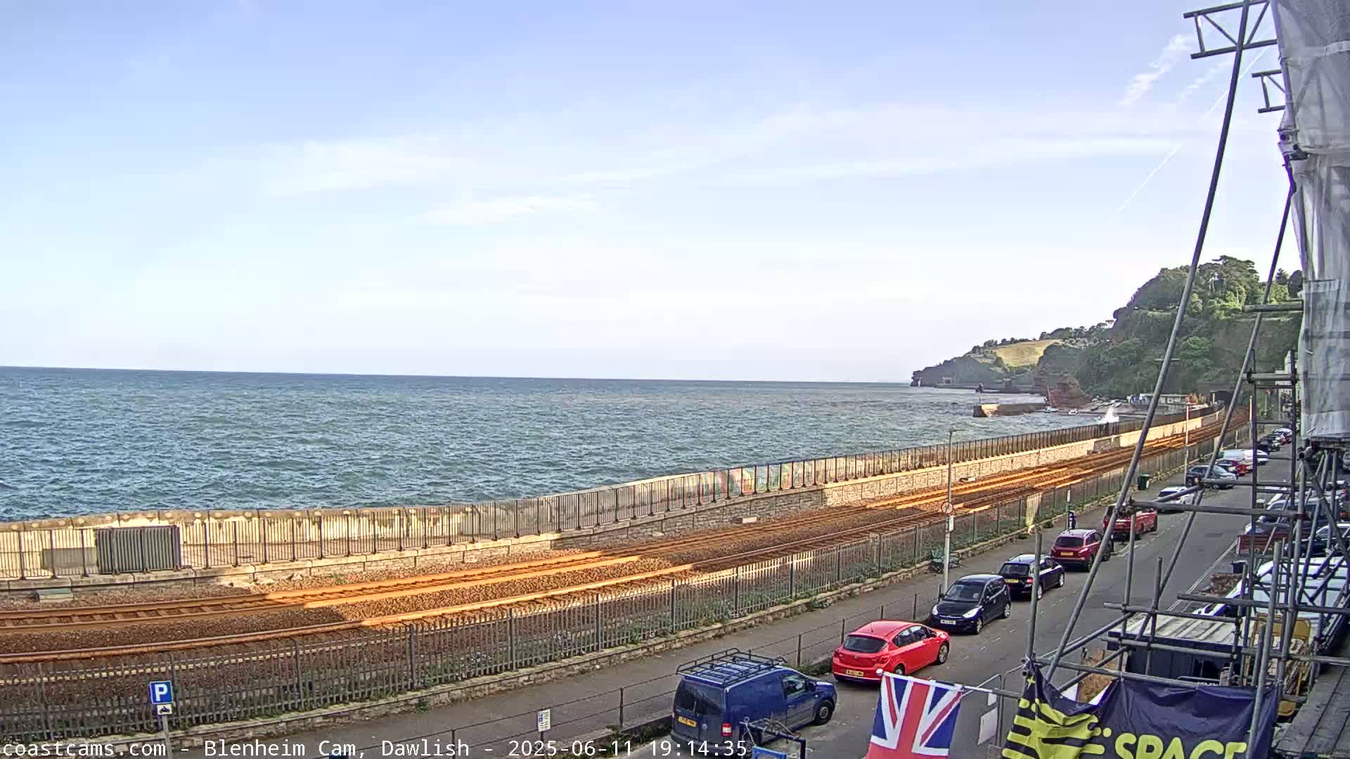 A seaside view shows train tracks running alongside a sea wall, with cars parked along a street next to the tracks under a mostly clear sky.