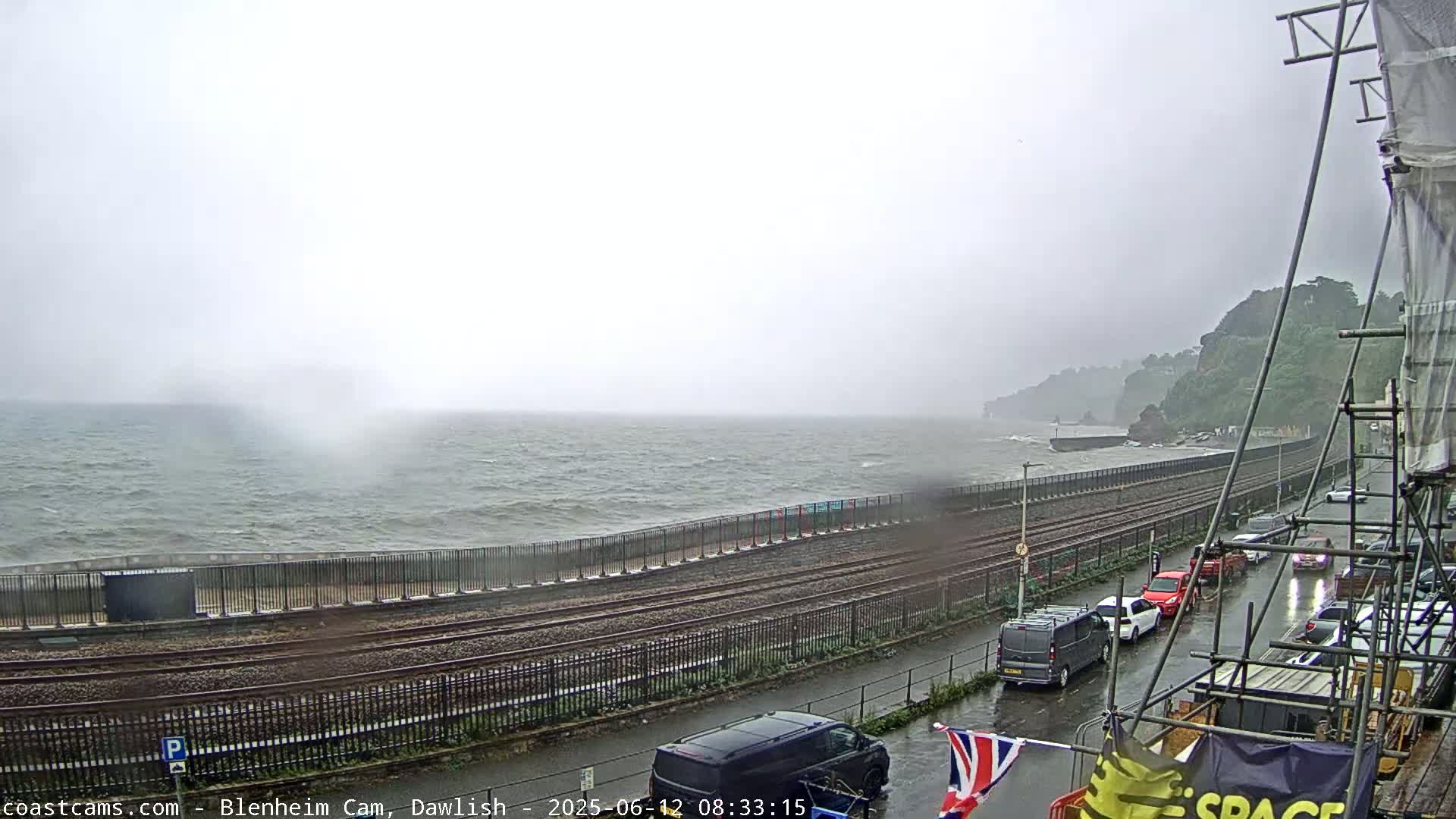 A stormy seaside scene shows a choppy sea with waves crashing against a seawall next to train tracks and a wet street with parked cars.