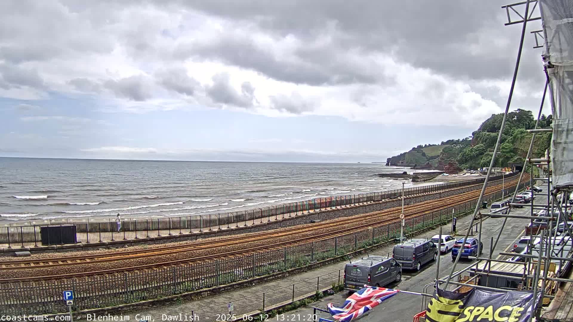 A coastal scene under cloudy skies shows a beach with gentle waves, railway tracks running alongside it, a road with parked cars, and a partially-constructed building.